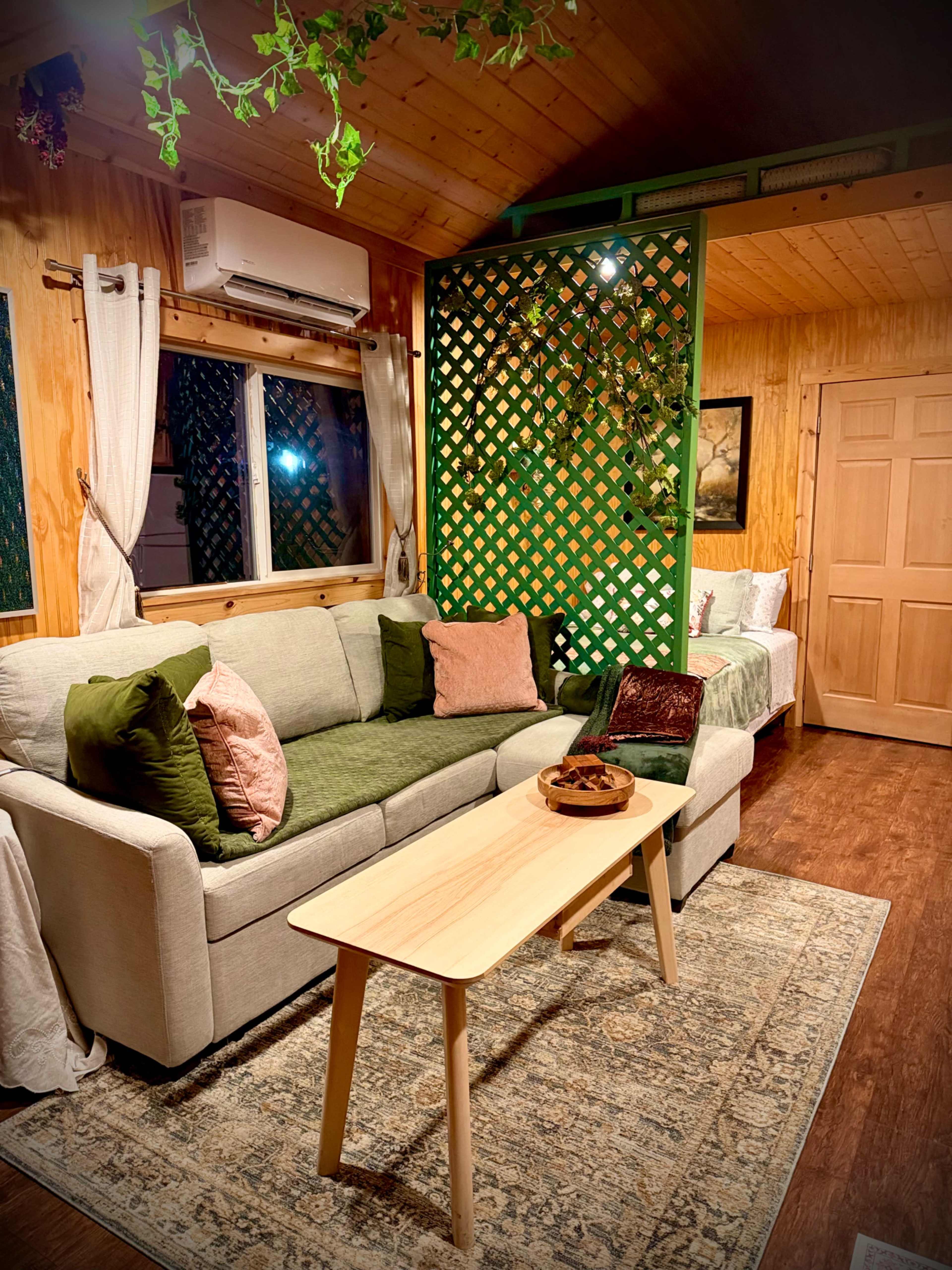 The cozy interior features a light-colored sofa with cushions, a wooden coffee table, and a partition with green latticework, all set in a warm-toned wooden room.