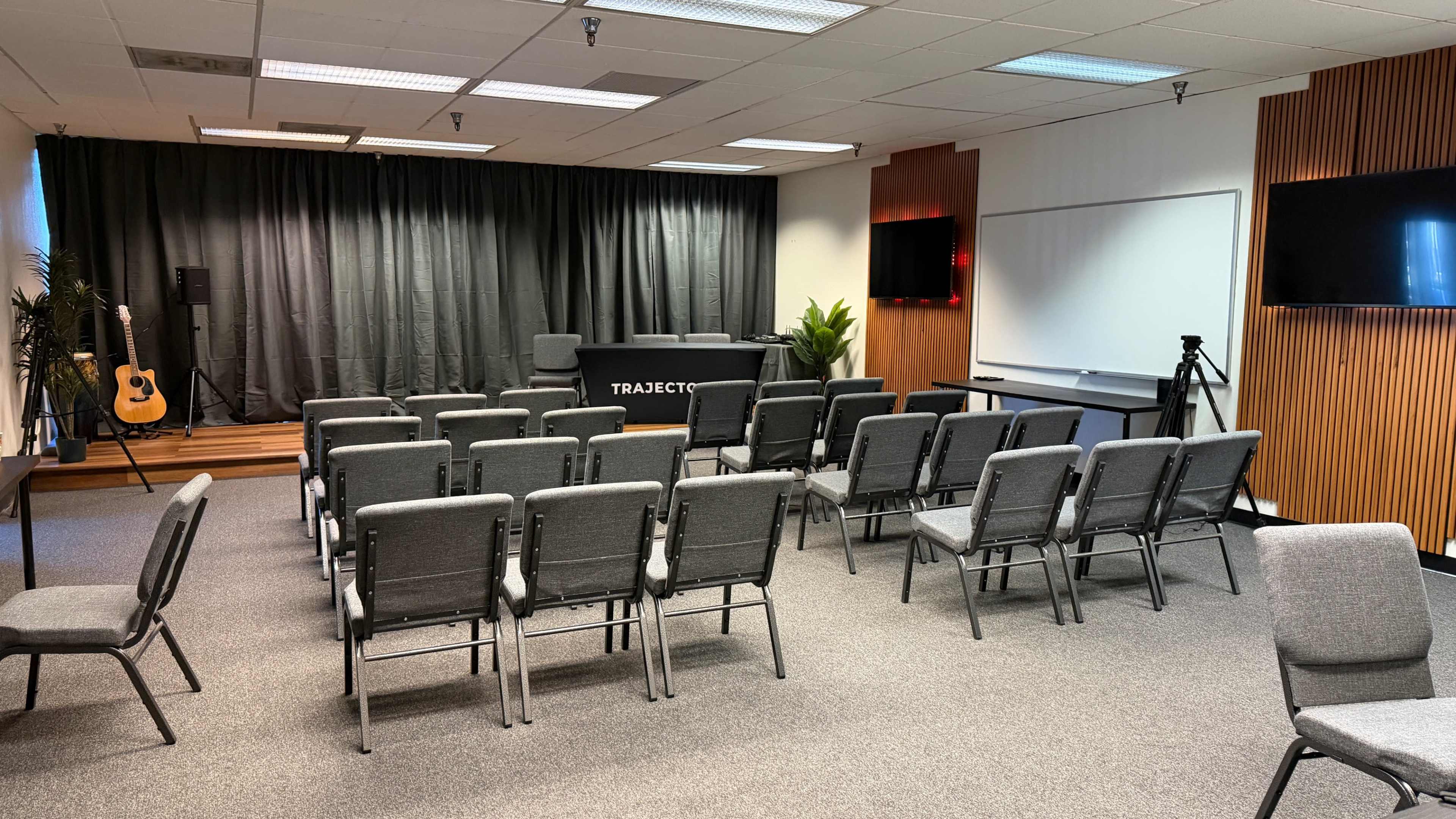 The image shows a conference room set up with rows of gray chairs facing a table, with a black curtain in the background and screens on the walls.