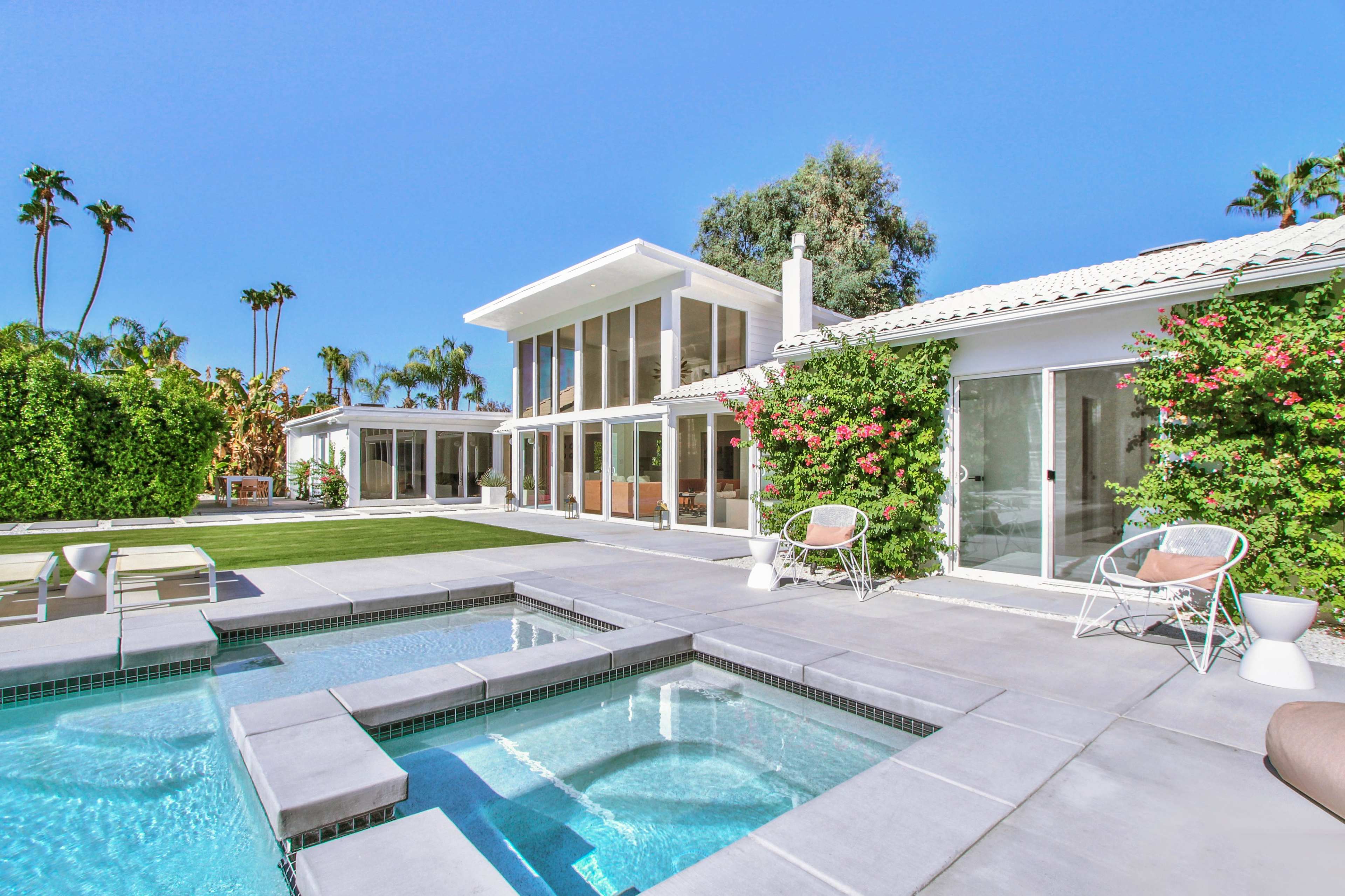 A modern house features a swimming pool and spa, surrounded by lush landscaping and bright blue skies.