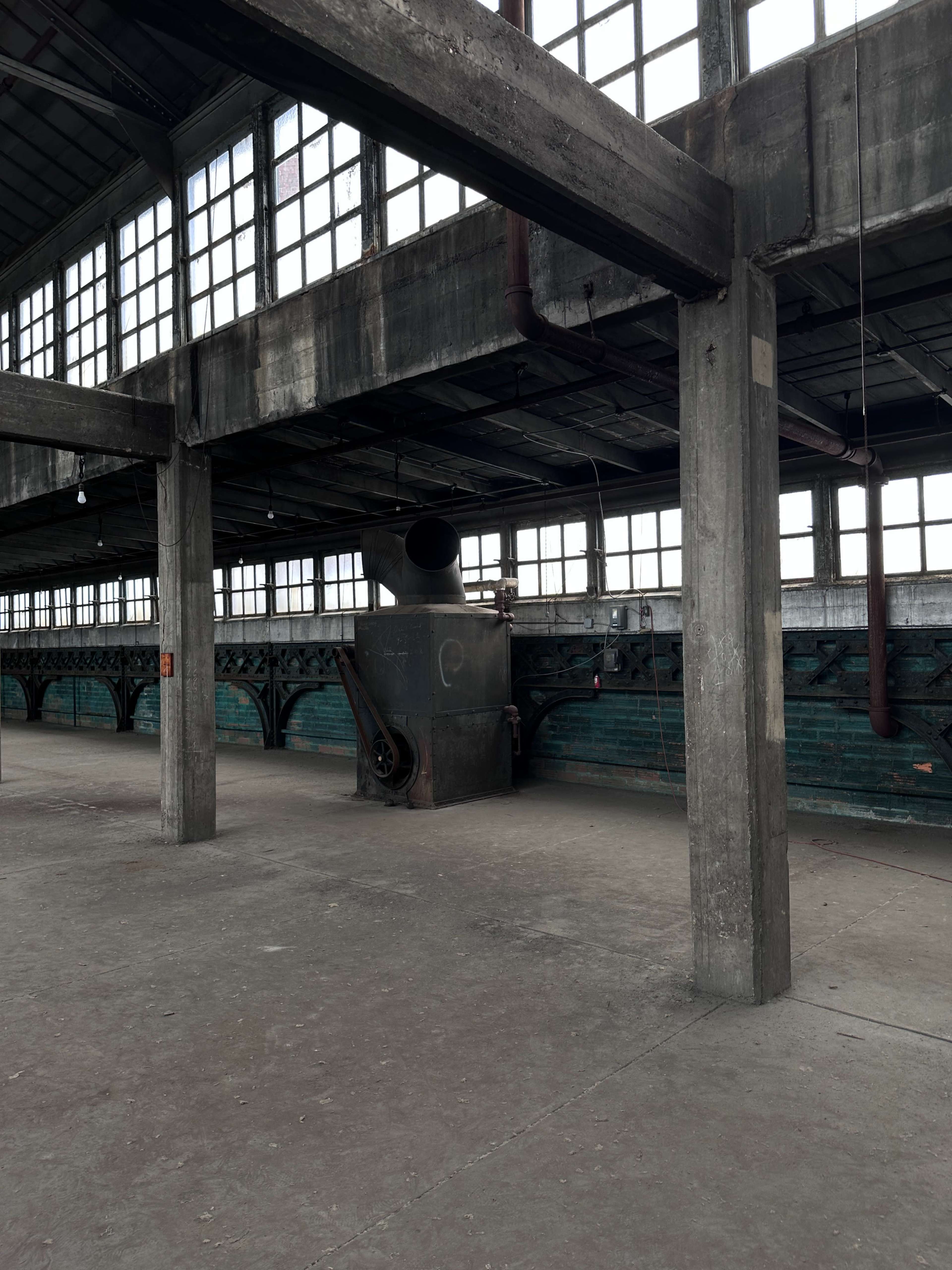 The image shows a large, empty industrial space with tall windows, exposed concrete pillars, and a metallic structure in the center.