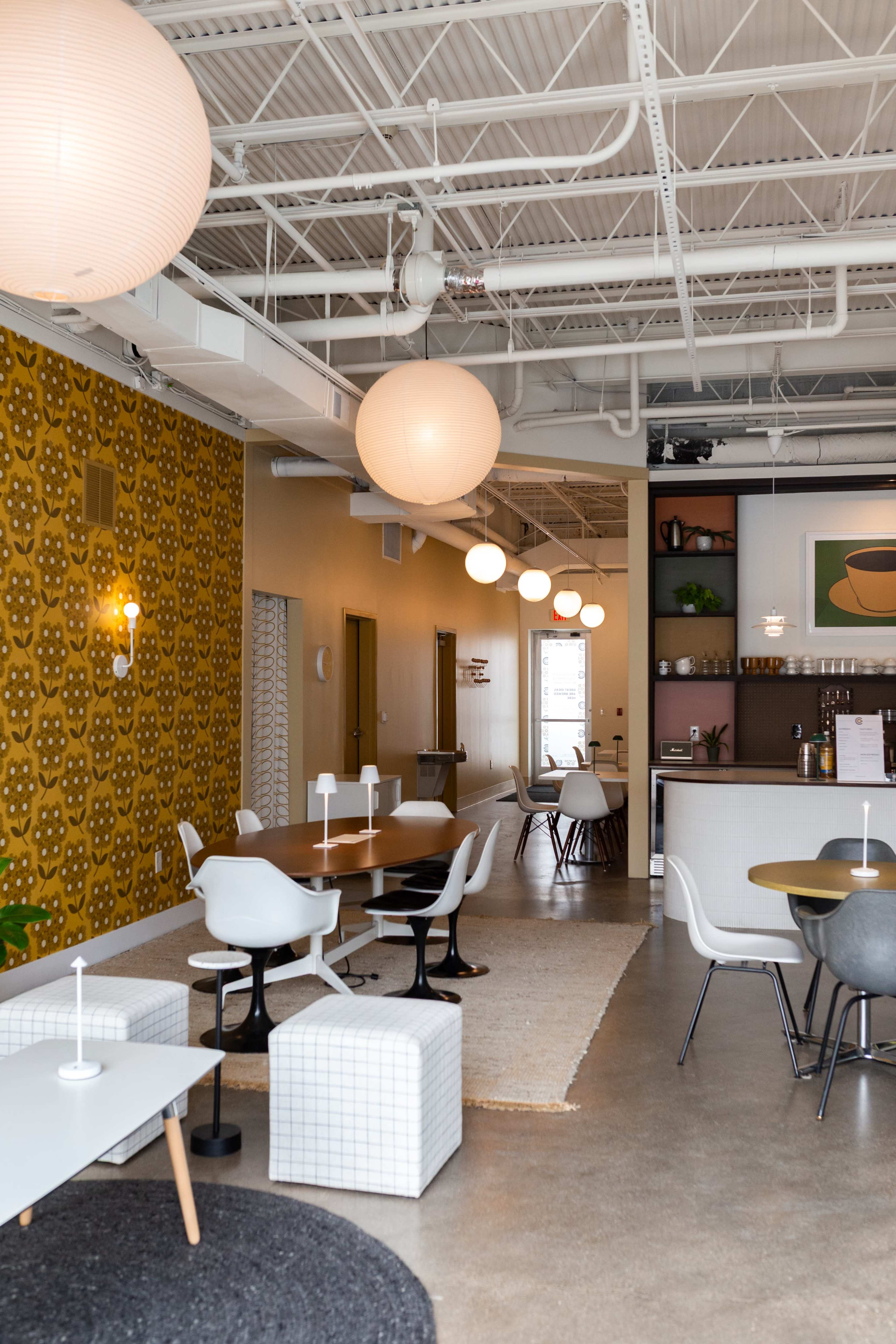 The image shows a modern café interior featuring various seating arrangements, pendant lighting, and a mix of tables and chairs against a patterned wall.