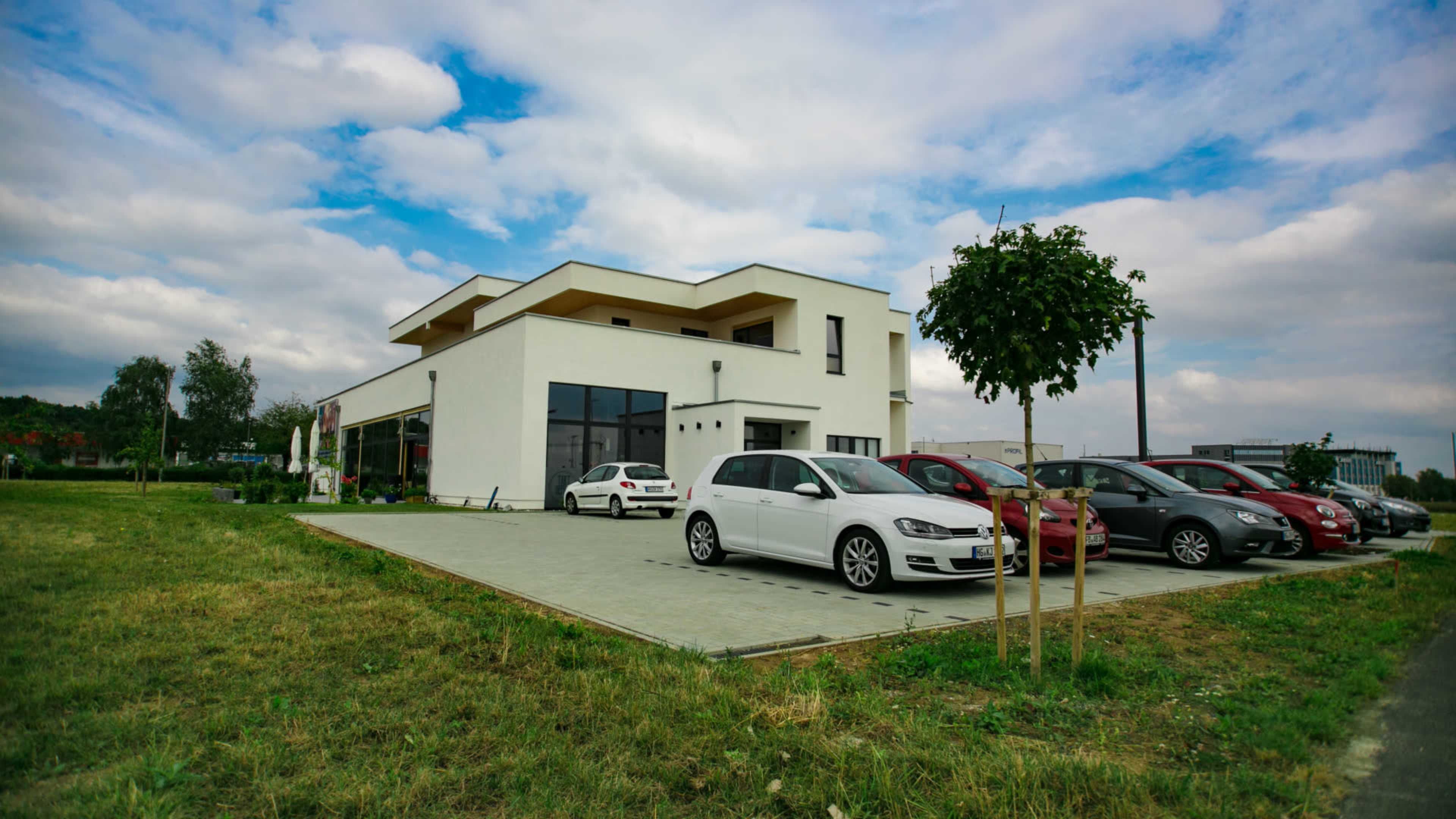 A modern white building is located beside a grass field, with several cars parked in front of it.