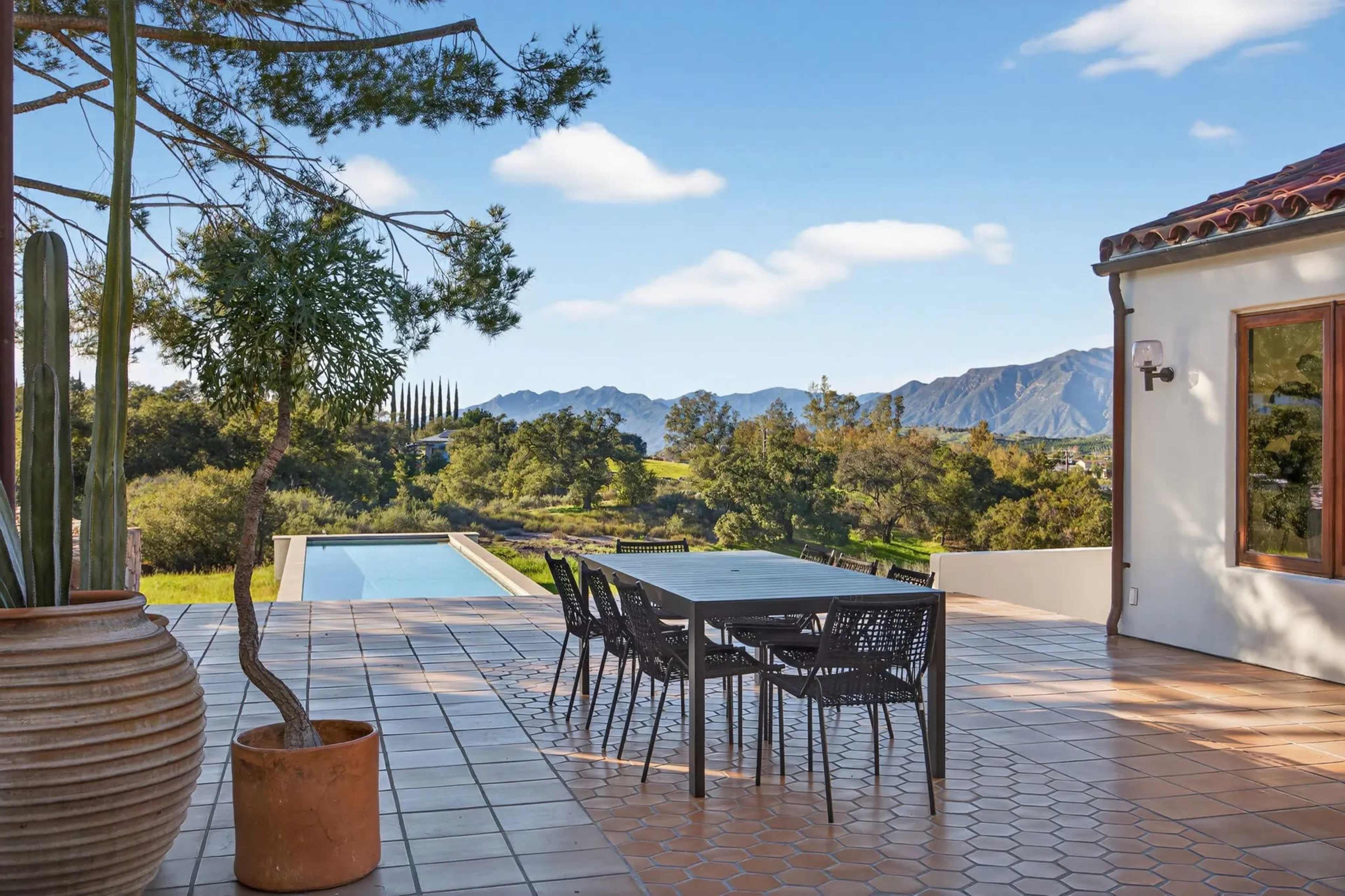A modern patio features a dining table and chairs overlooking a swimming pool and a mountainous landscape.