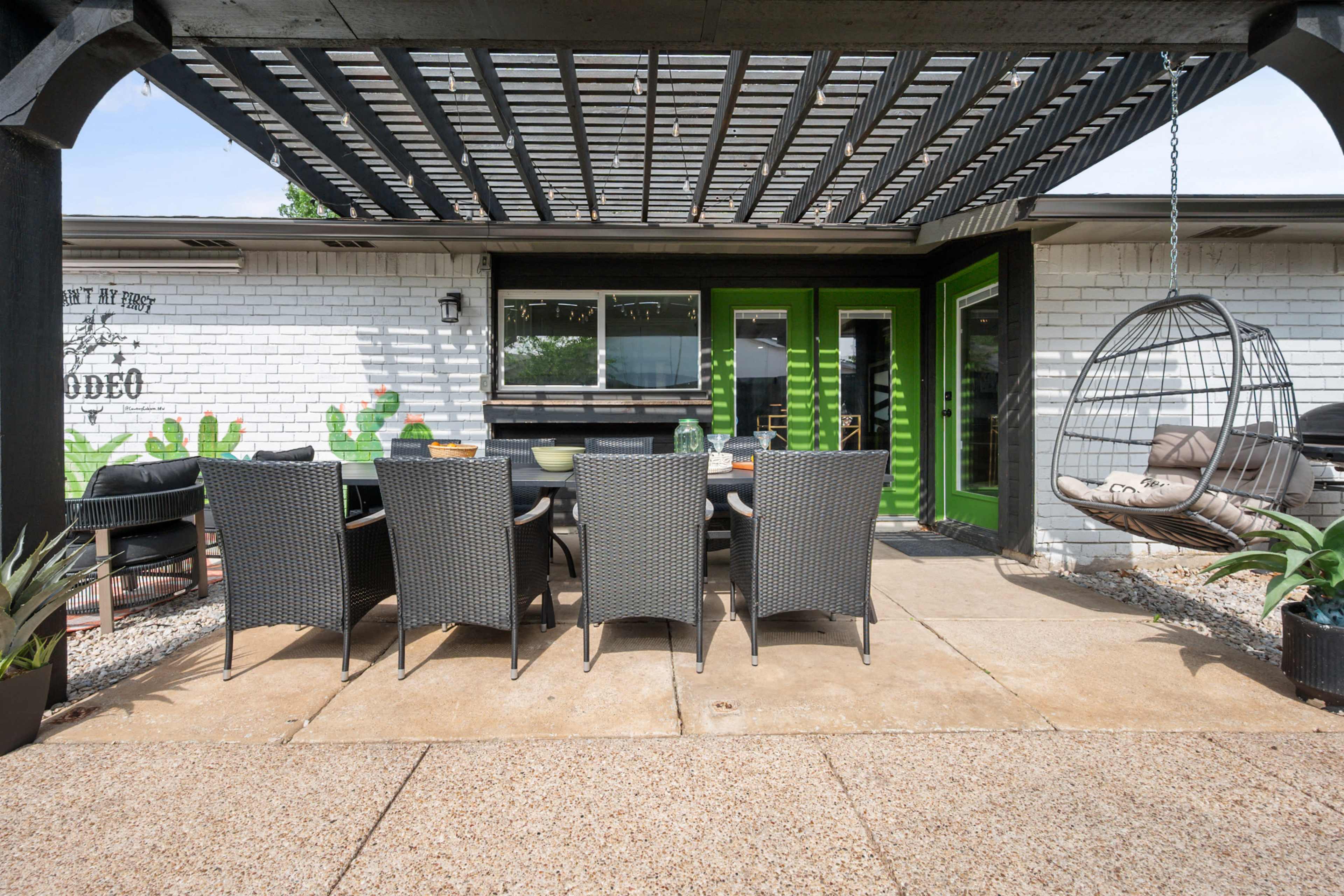 A patio features a long dining table with wicker chairs under a pergola, adjacent to a house with green doors and decorative cactus artwork on the wall.