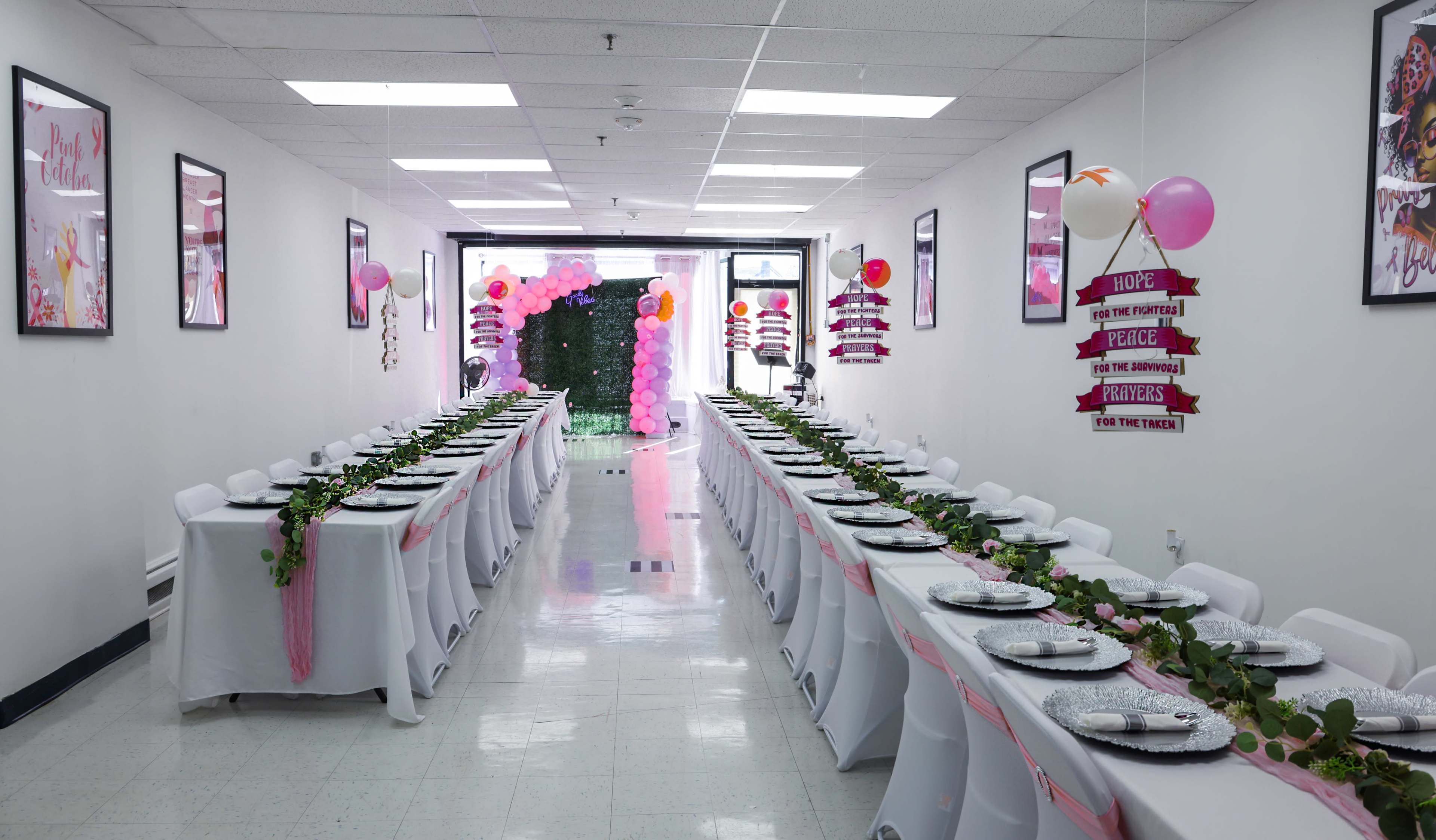 A long, decorated hallway features rows of tables set for a dining event, adorned with pink table runners and floral centerpieces, while decorative balloons and signs enhance the festive atmosphere.