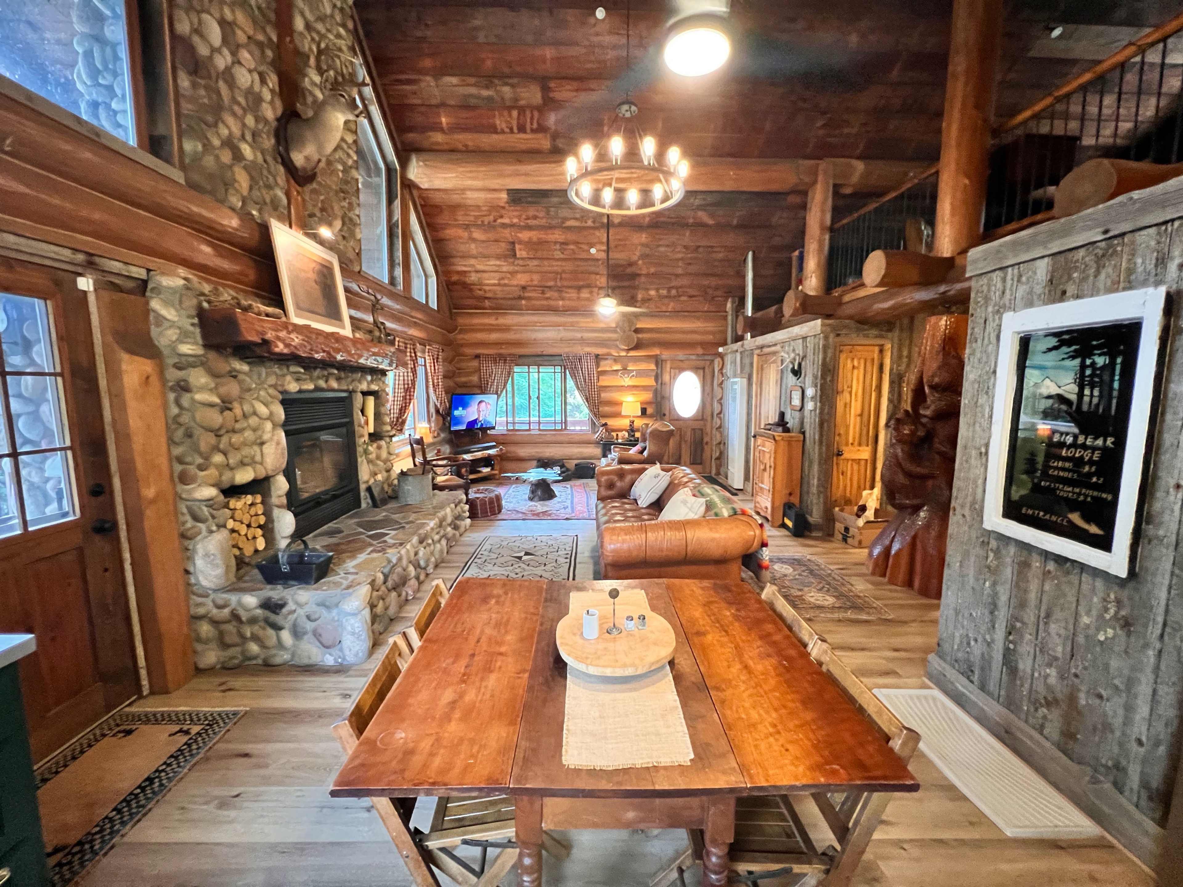 The image shows a rustic cabin interior featuring a wooden table in the foreground, a stone fireplace, and furniture arranged around a cozy living space.