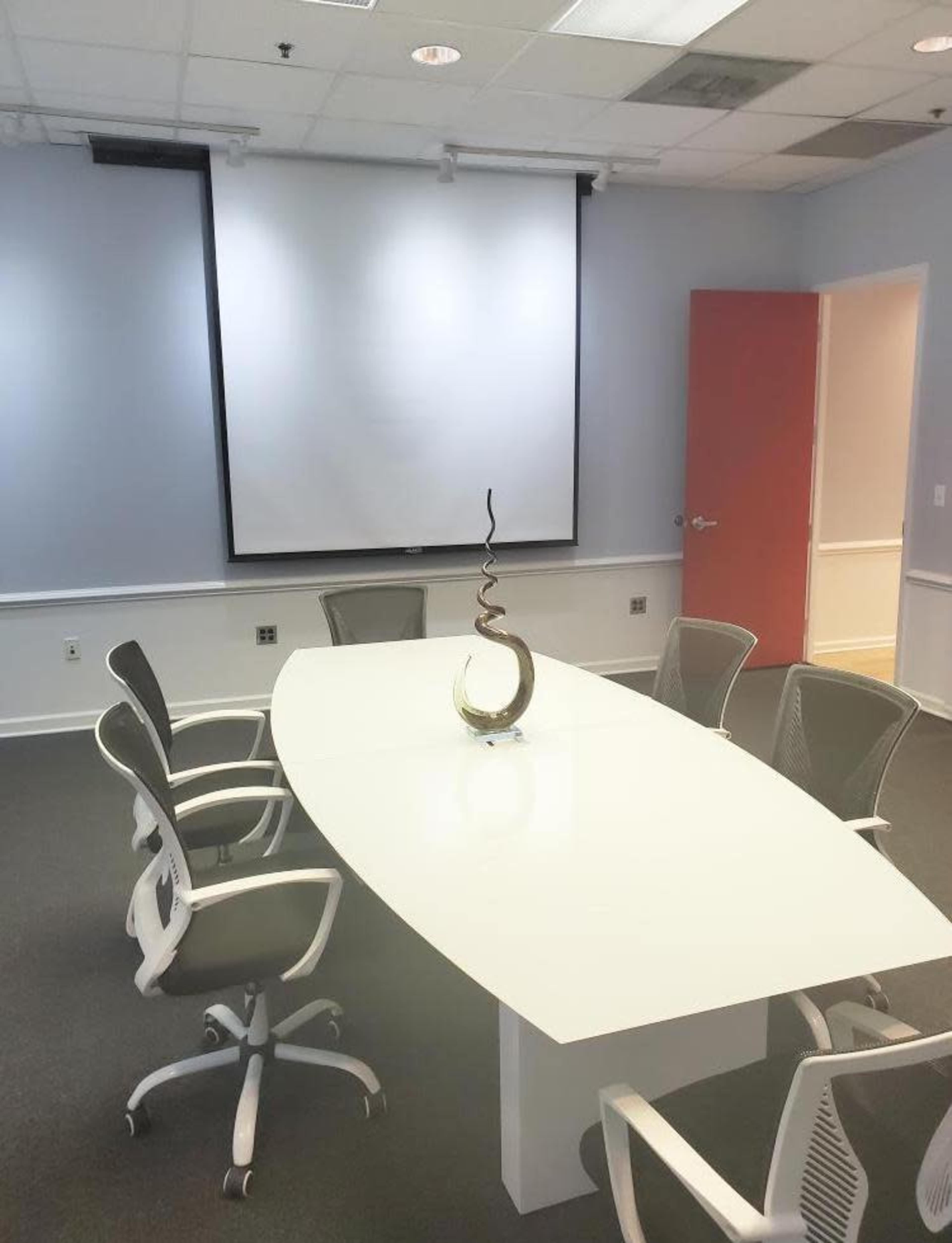A modern conference room with a white oval table, five ergonomic chairs, and a decorative sculpture in the center, along with a large projection screen on the wall.