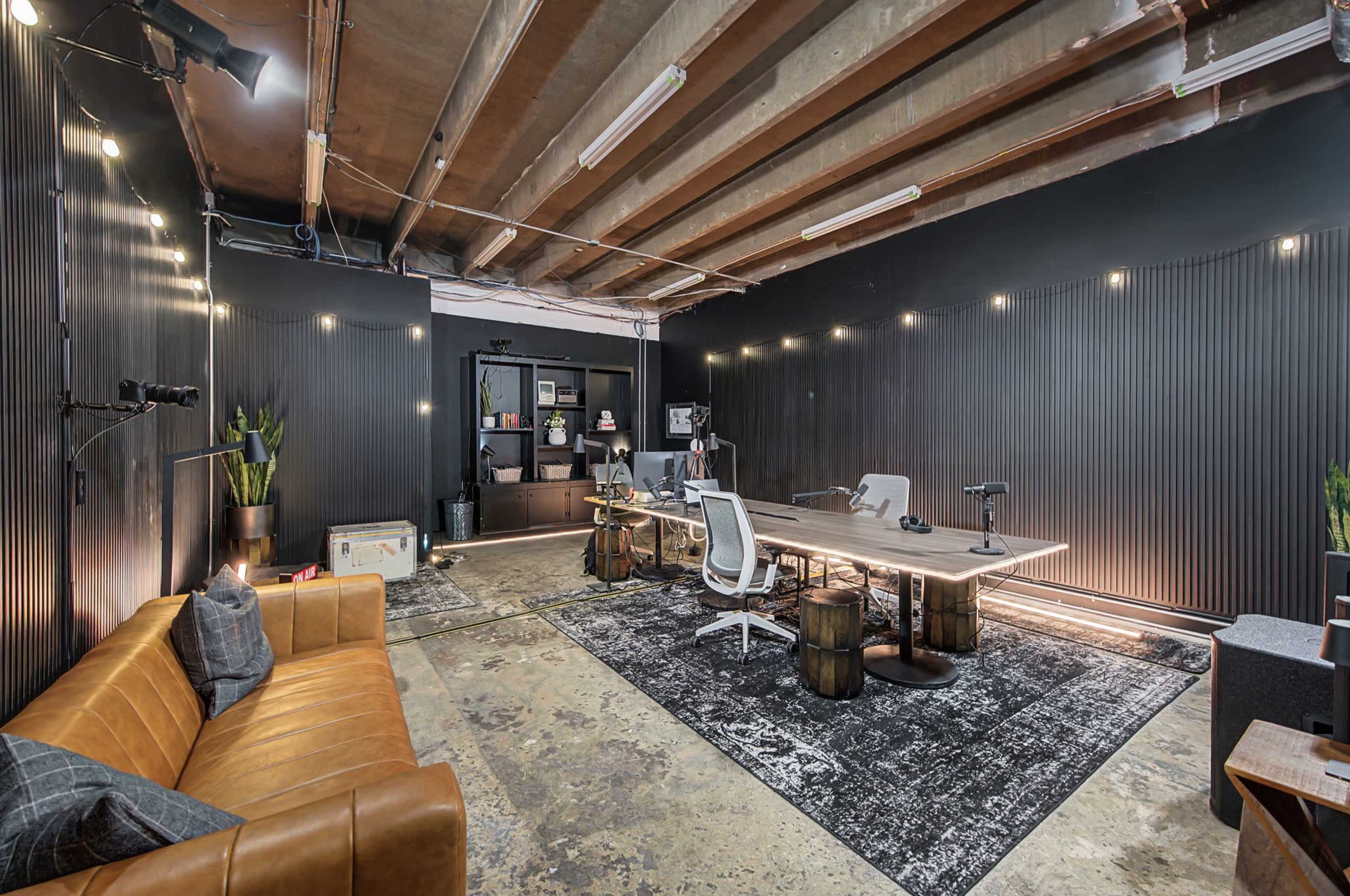 The image shows a modern office space with a brown leather sofa, a large conference table, and task lighting against a black wall with wooden paneling.