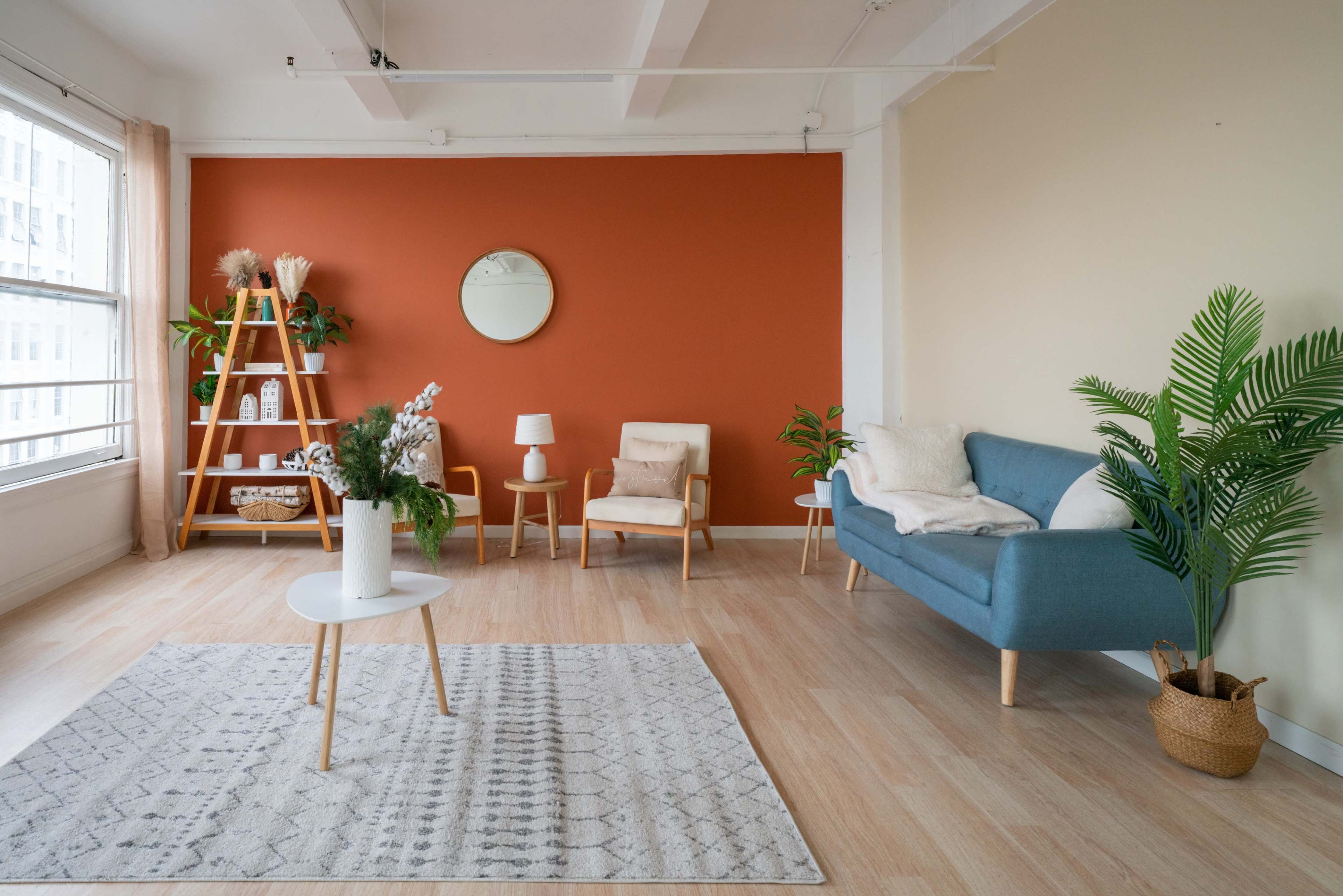 The image shows a modern living room with a blue sofa, a small white table, a patterned rug, and plants, alongside an orange accent wall and a round mirror.