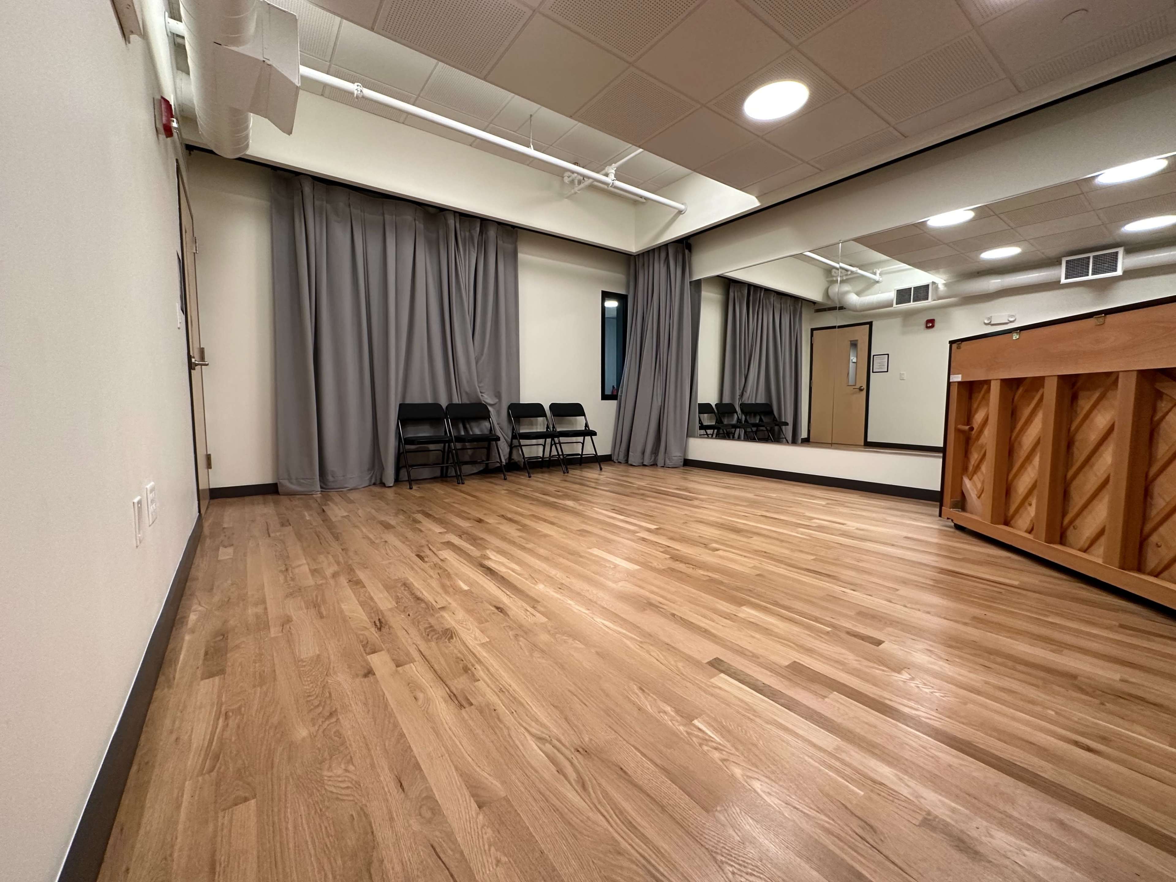A spacious room with wooden flooring, gray curtains, a mirror, and several black chairs lined up along one wall.