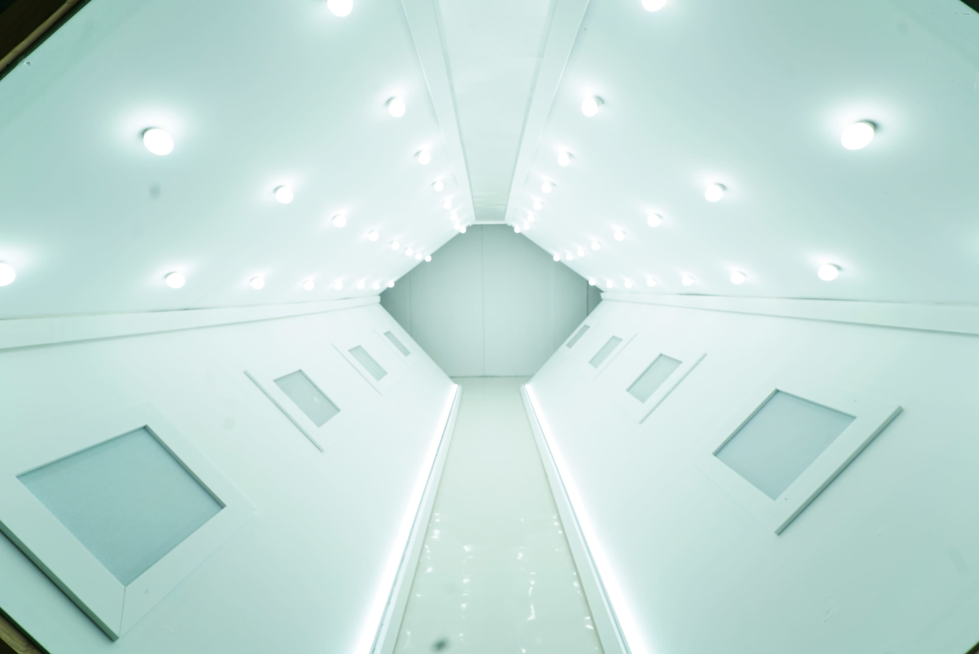 The image shows a long, narrow hallway with white walls and a series of bright lights overhead.