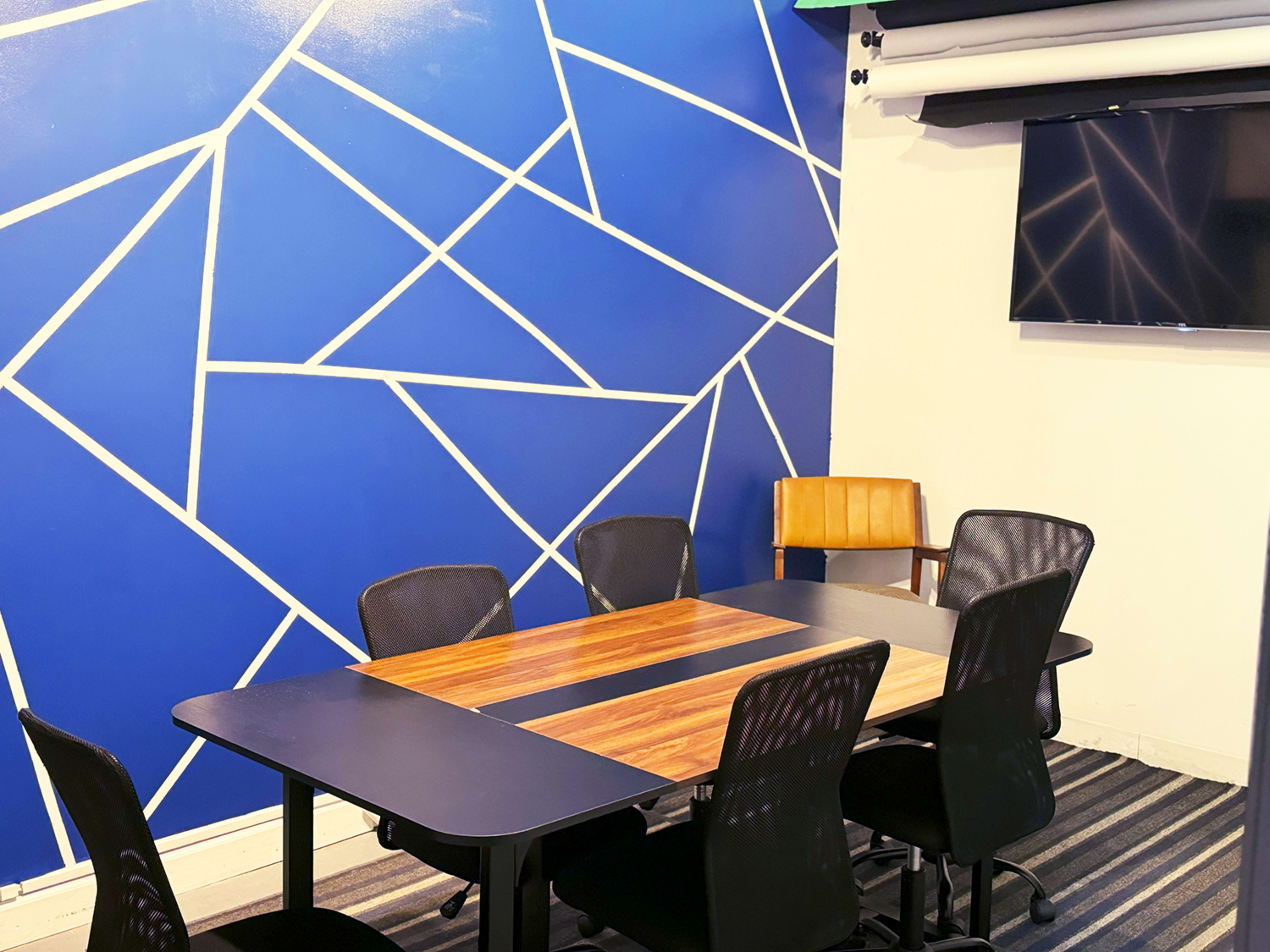A conference room features a wooden table surrounded by six black mesh chairs, with a blue geometric wall design and a mounted television.