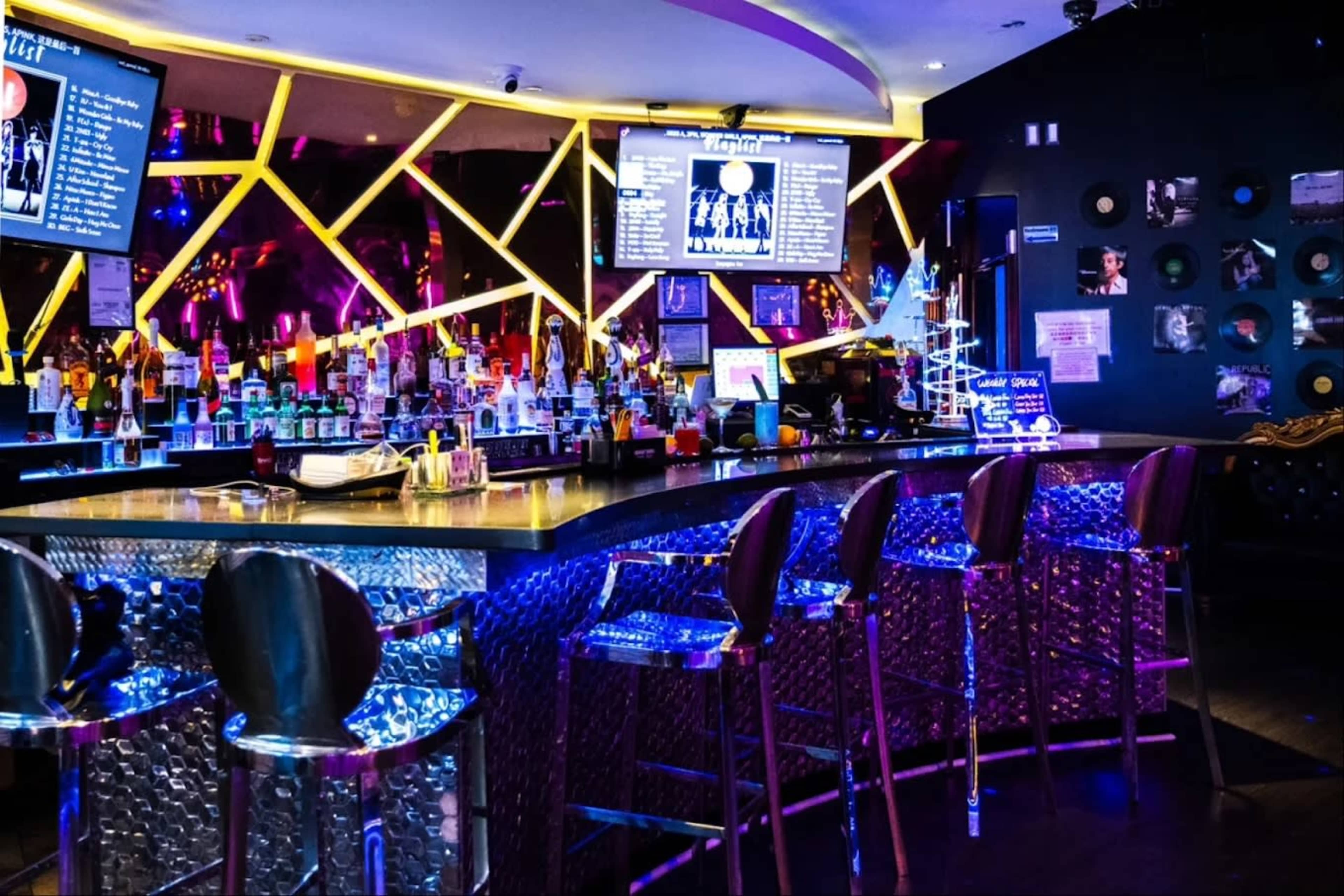 The image displays a vibrant nightclub bar with colorful lighting, a well-stocked bar counter, and a row of metallic bar stools.