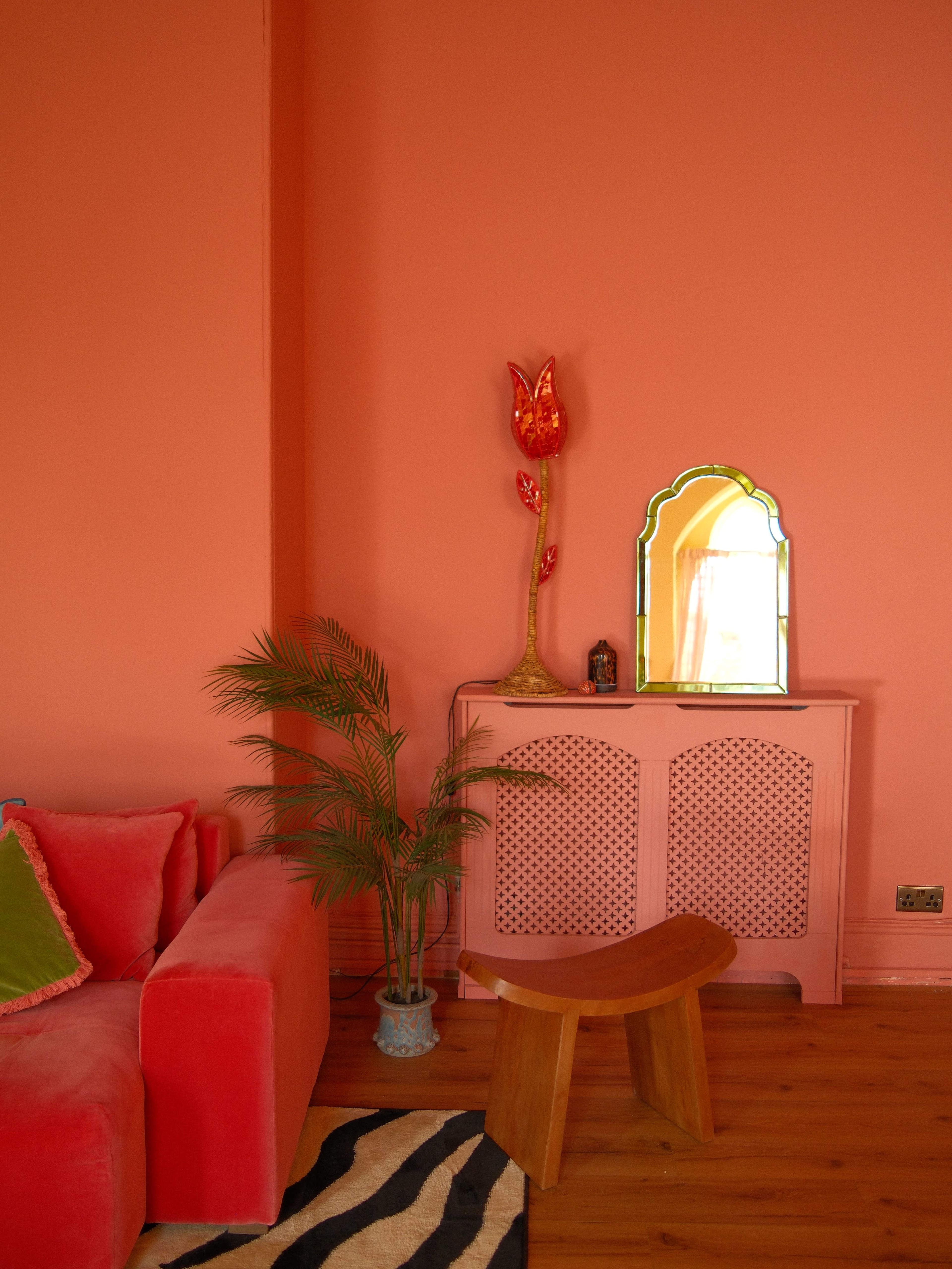 The image shows a vibrant room with coral-painted walls, a coral sofa, a wooden stool, a decorative mirror, and a plant beside a colorful lamp.