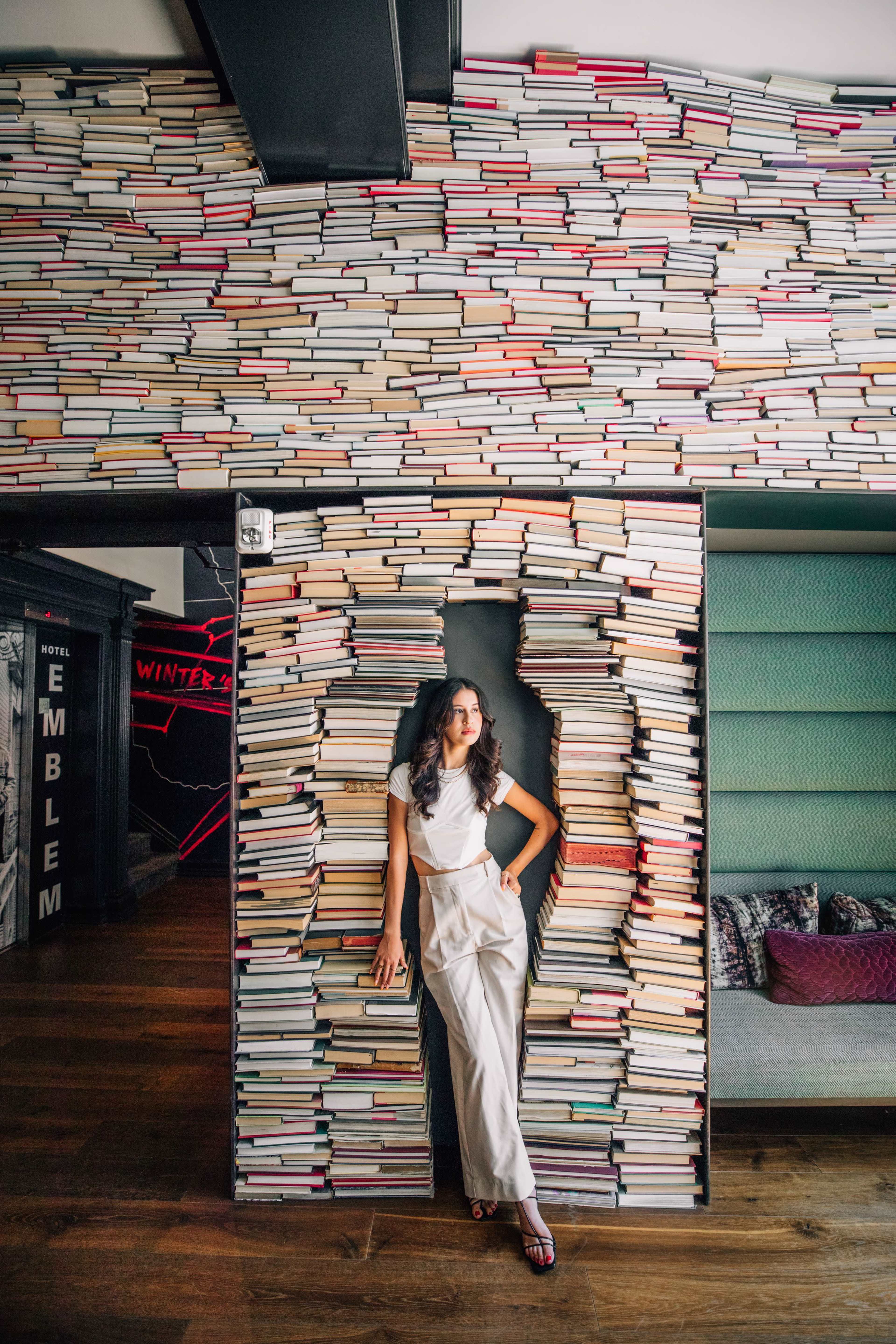 A woman in a stylish outfit stands in a doorway framed by stacked books on a wall.