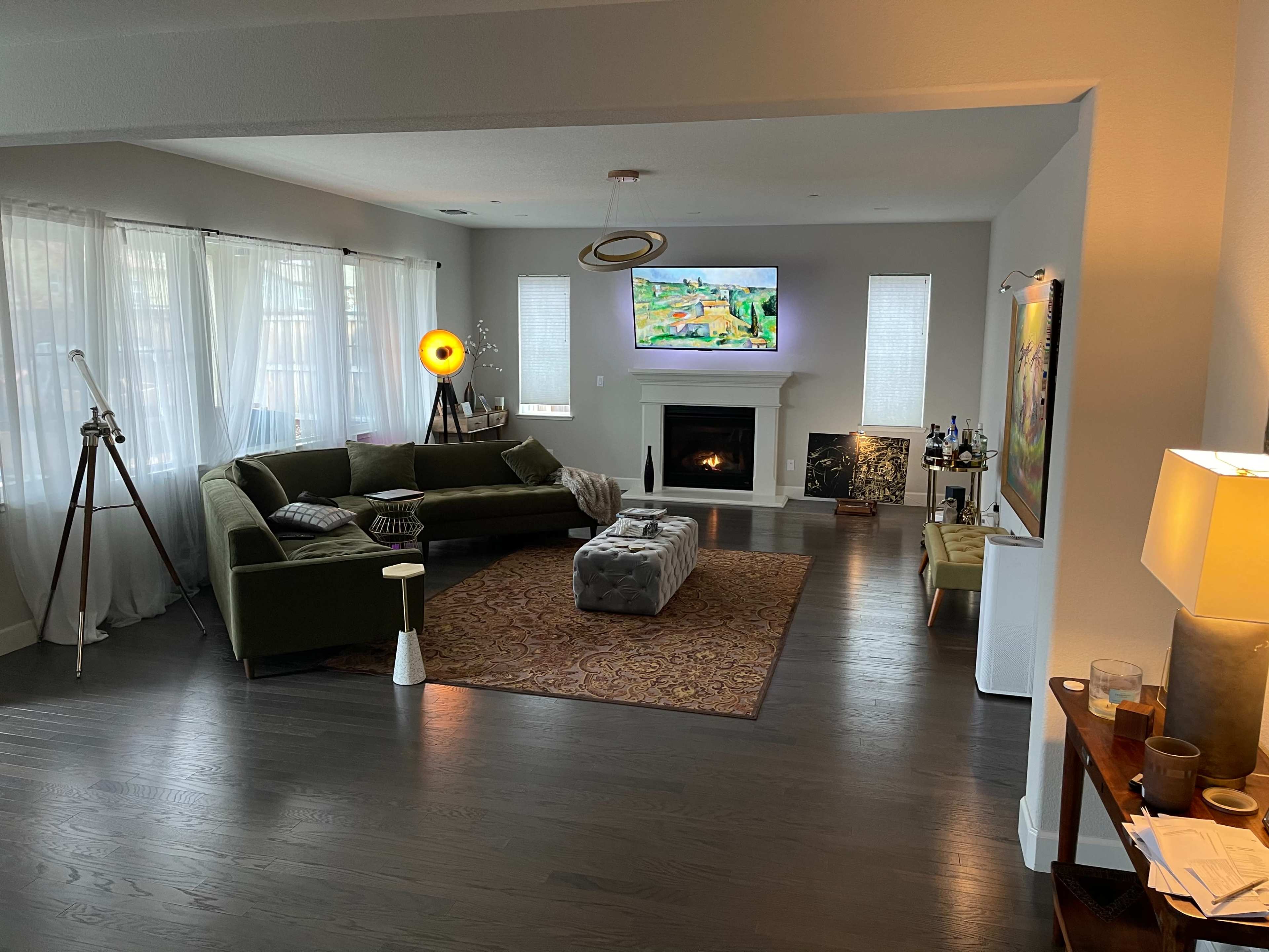 A modern living room featuring a green sectional sofa, an ottoman, a patterned area rug, a wall-mounted television, and large windows with light curtains.