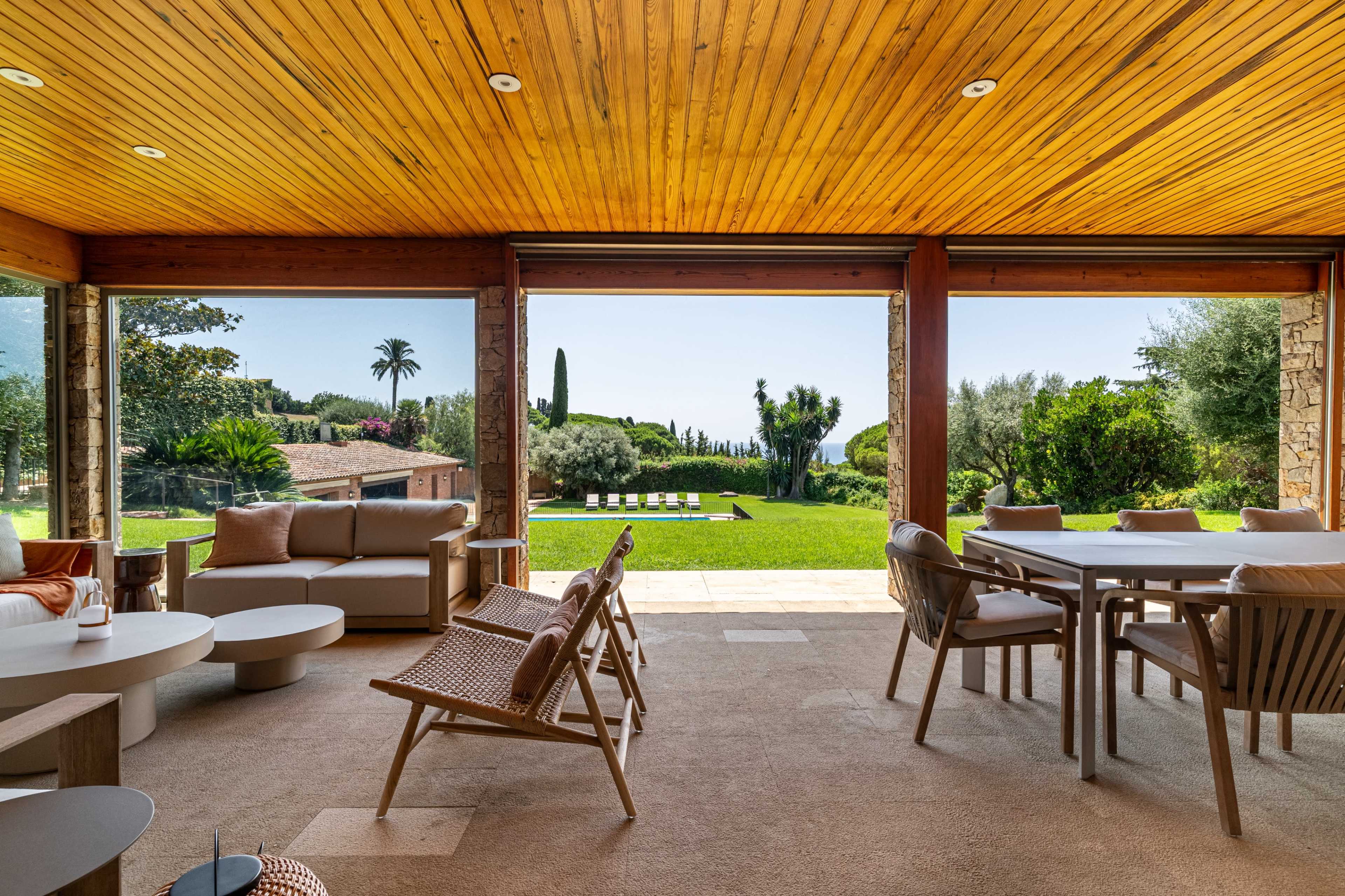The image shows a modern living space with wooden ceilings, cozy furniture, and large windows providing a view of a green outdoor landscape with trees and a pool.