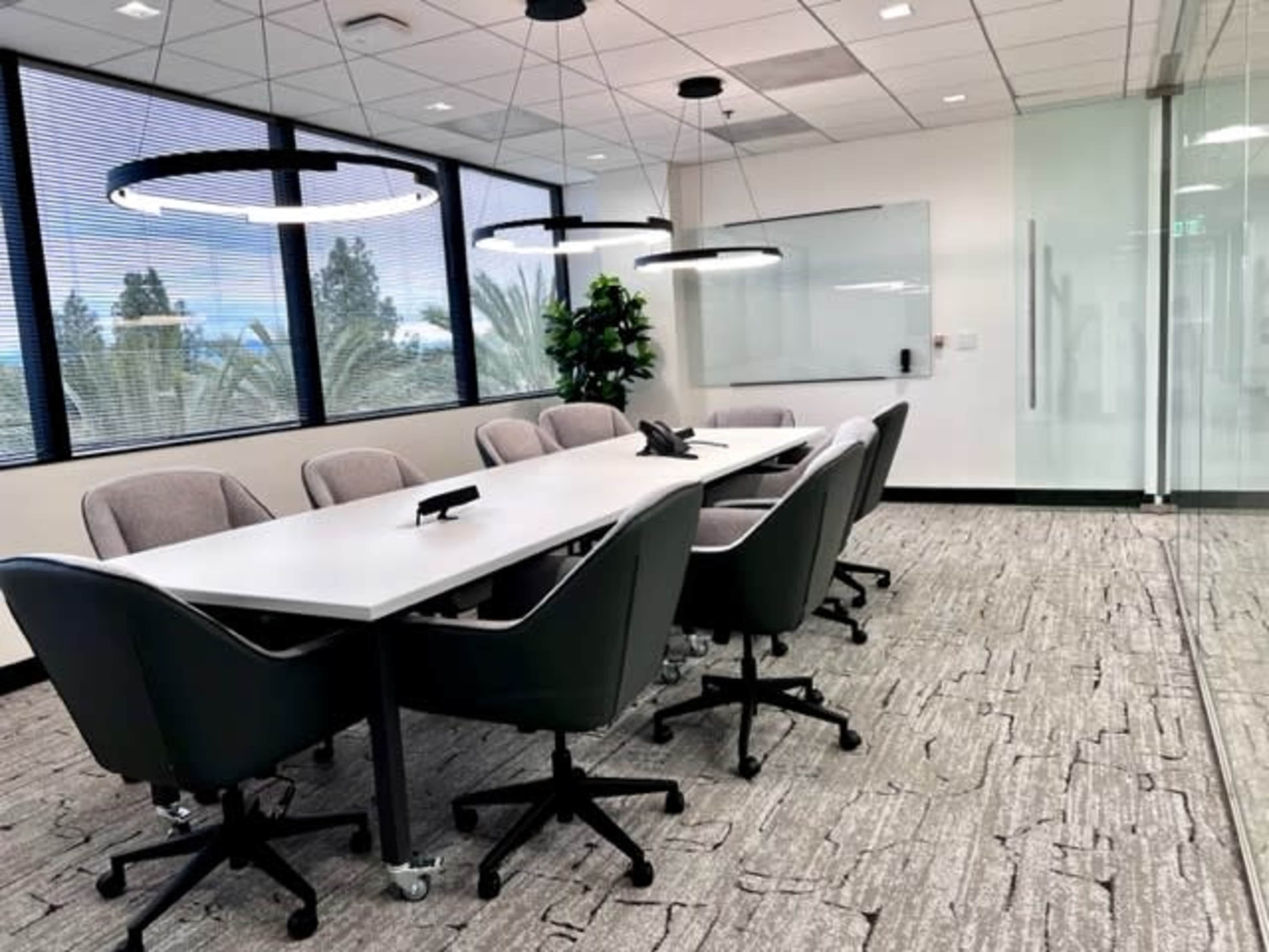 A modern conference room features a long table surrounded by ergonomic chairs and large windows with a view of greenery outside.