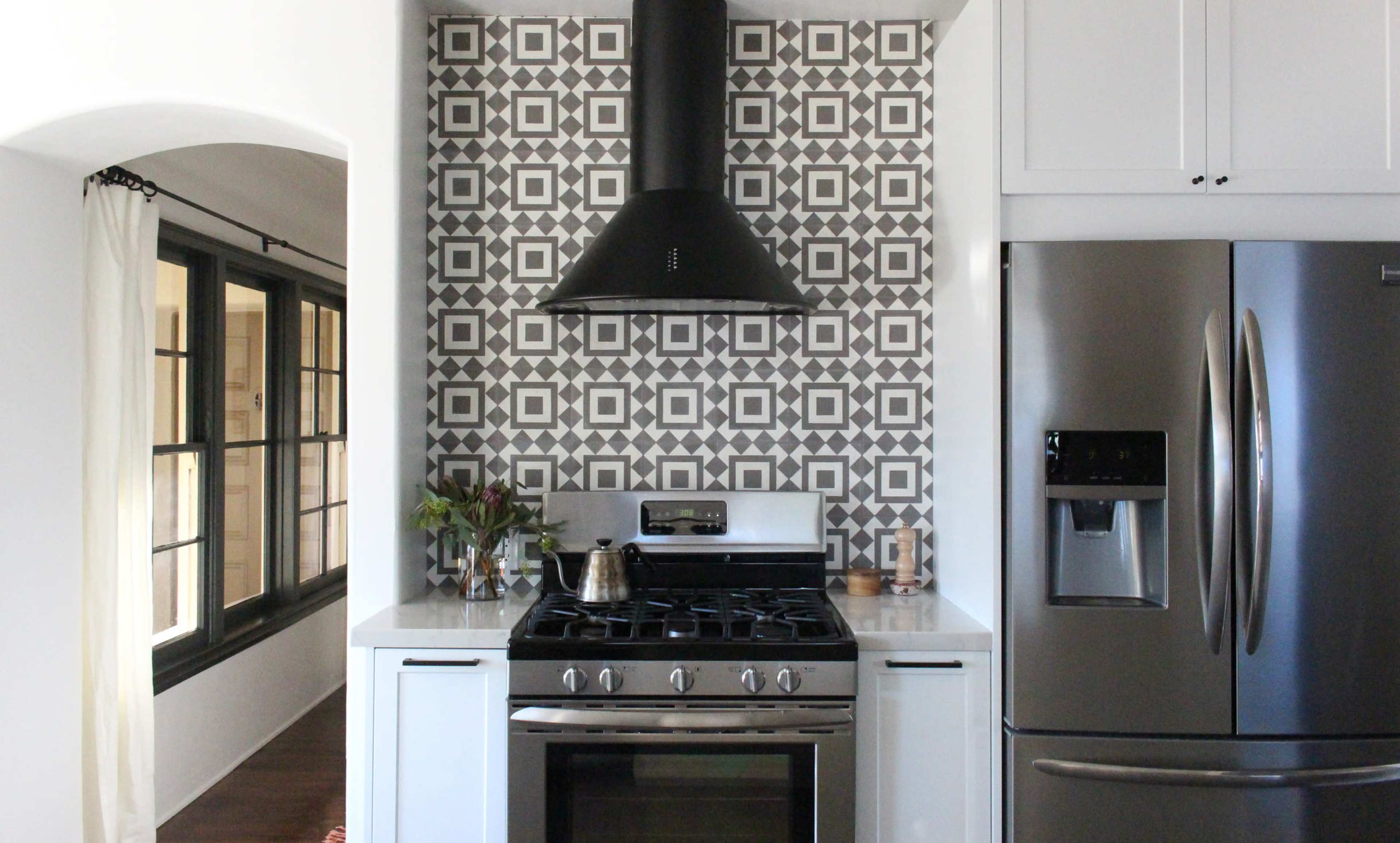 A modern kitchen featuring a stainless steel gas range, a large refrigerator, and a decorative tiled backsplash with a black exhaust hood.