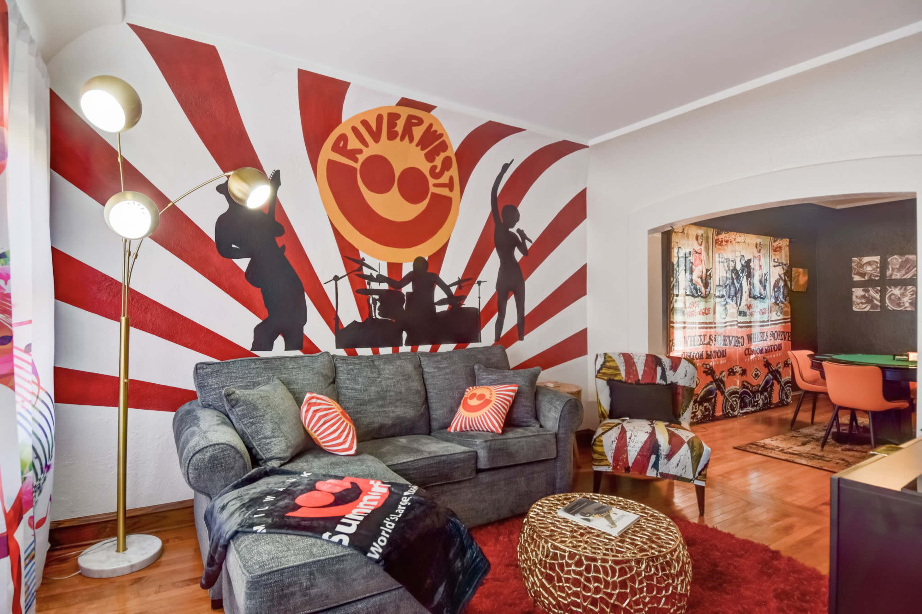 A living room with a gray couch, a floor lamp, a vibrant mural featuring silhouettes of musicians, and colorful decorative elements.