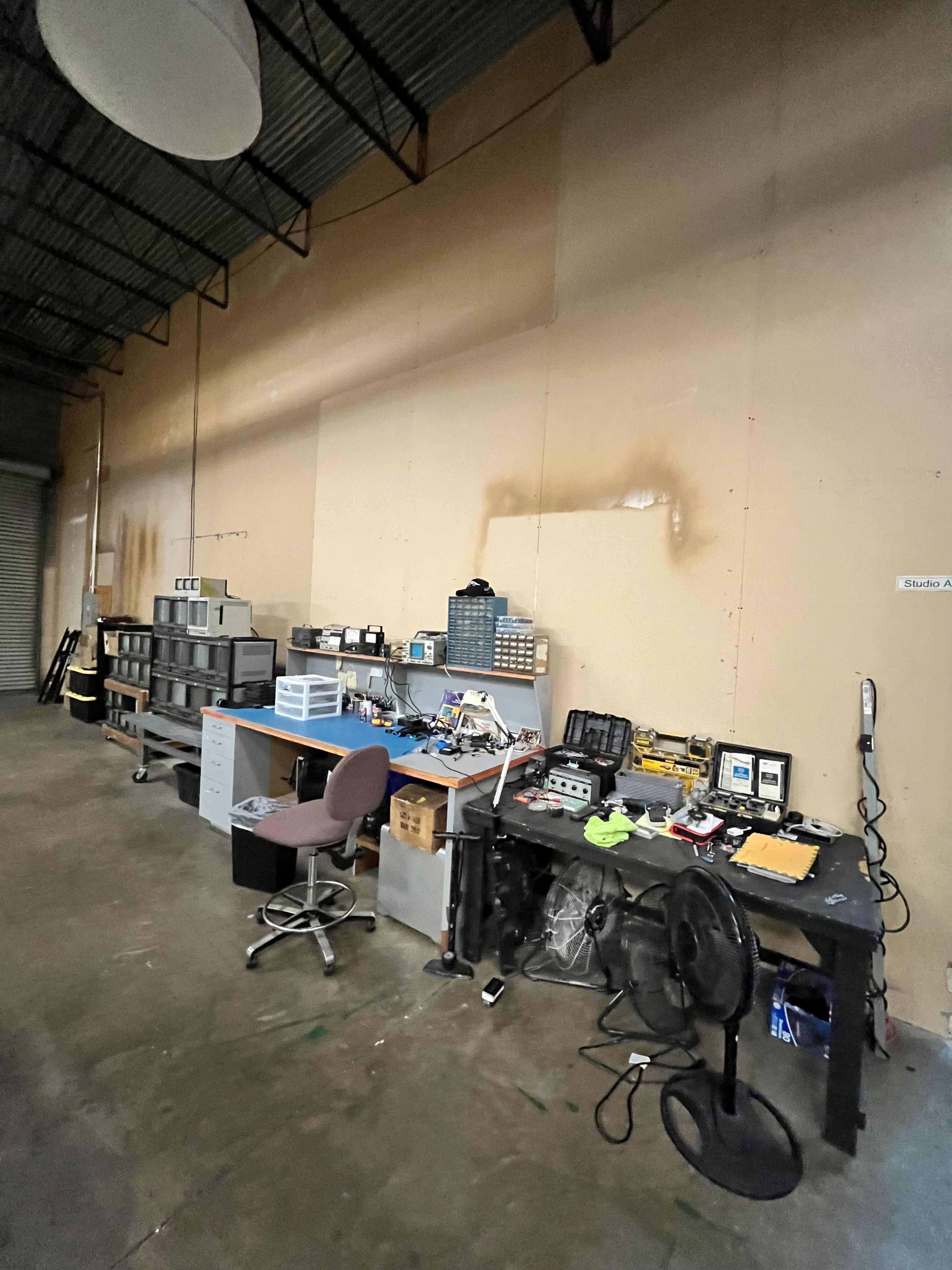 The image shows a spacious workspace with various tables, storage bins, and electronic equipment arranged against a plain wall.