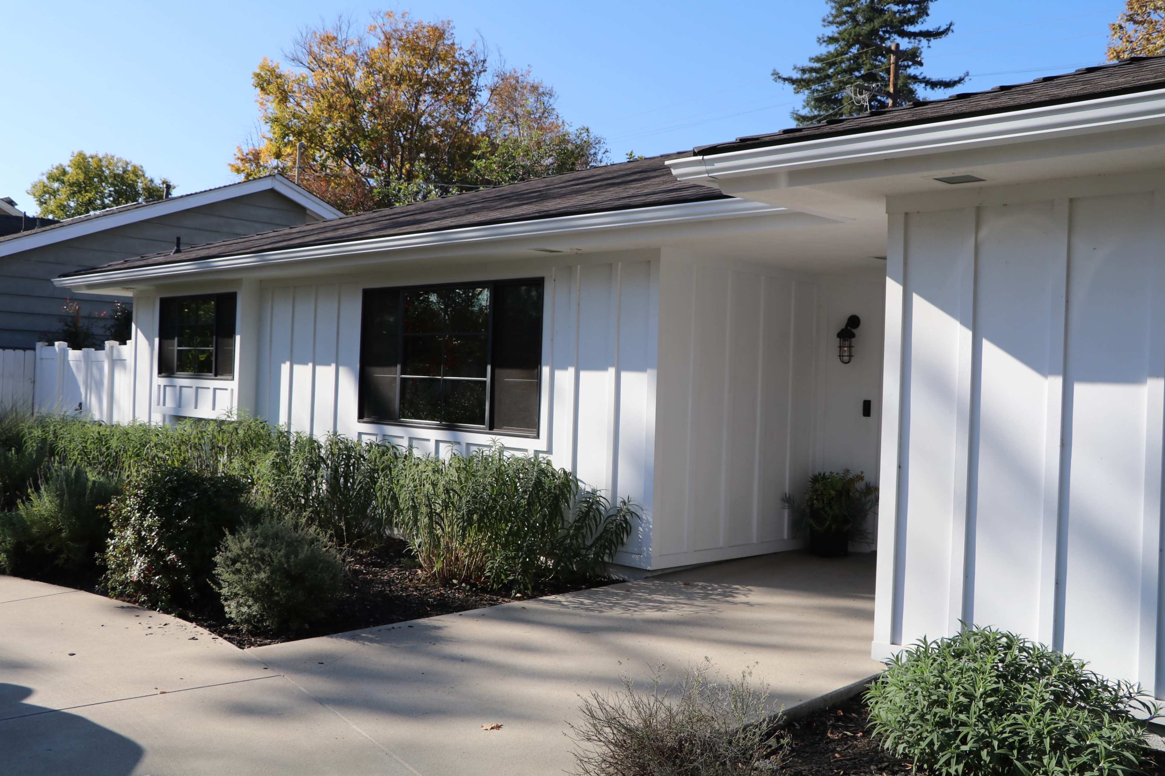 A modern, white-sided house with large windows and a concrete pathway surrounded by greenery.