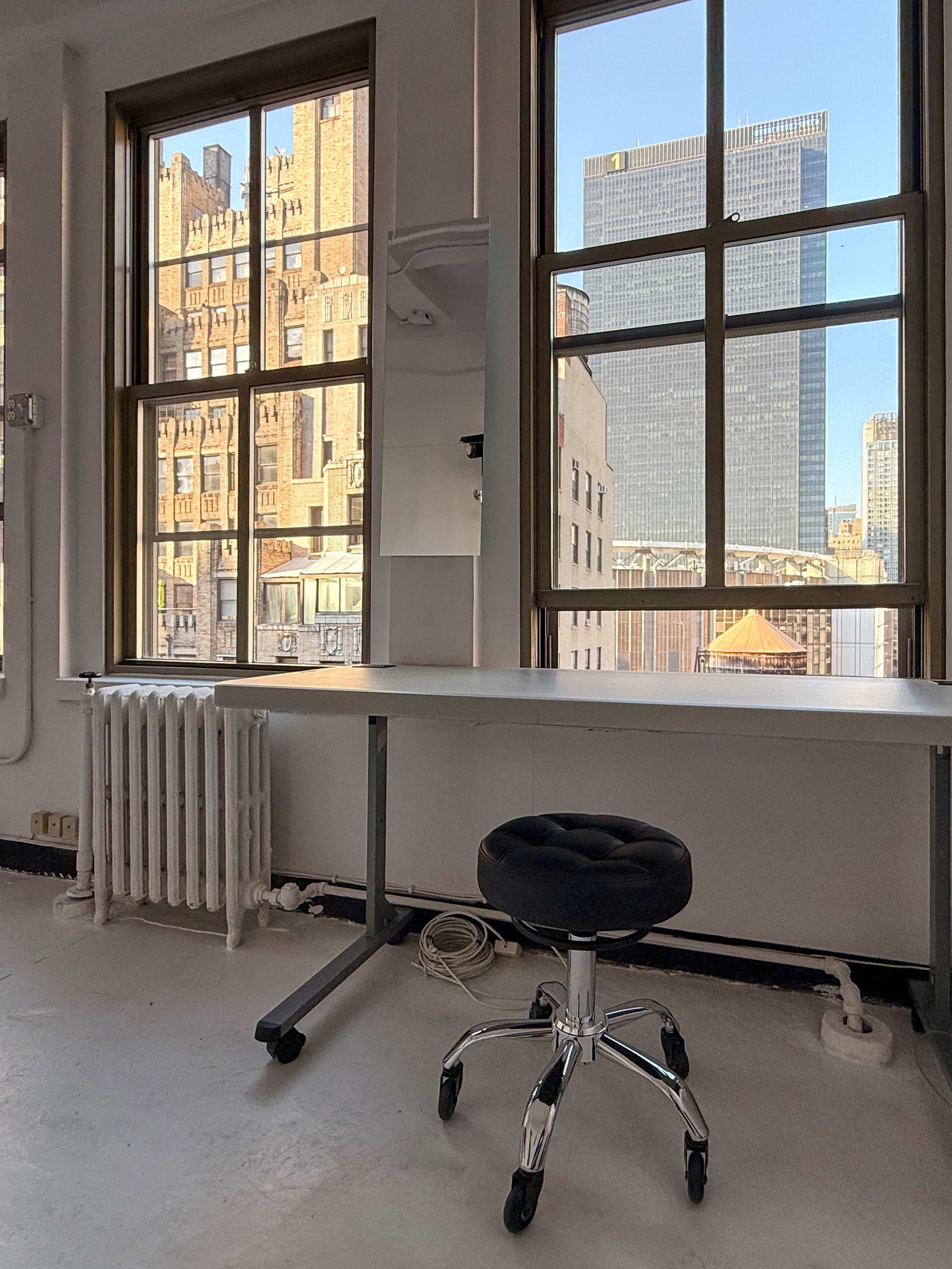 The image shows a simple workspace featuring a stool, a table, and large windows overlooking a cityscape with tall buildings.