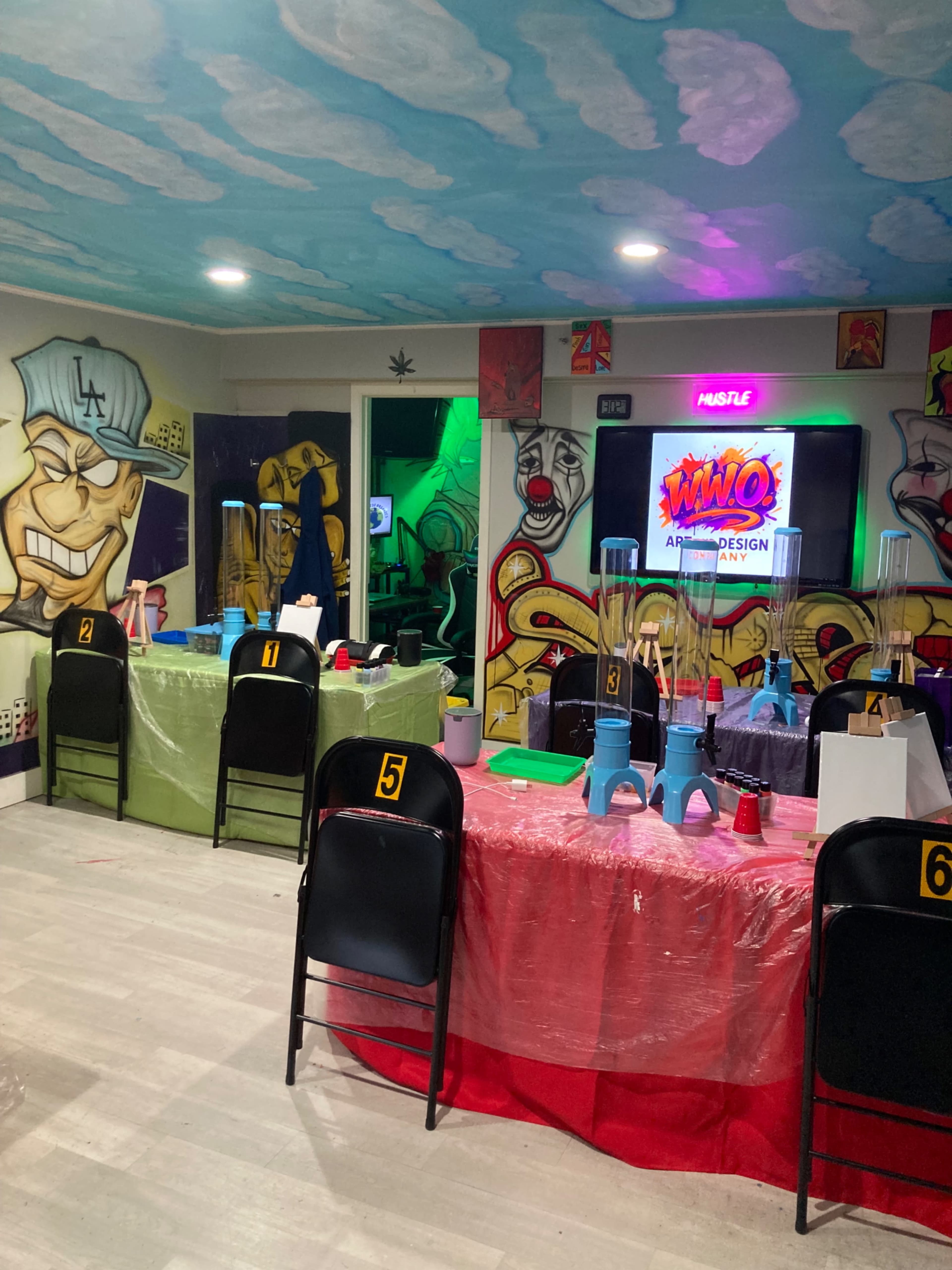 The image shows a vibrant room decorated with colorful graffiti murals, featuring tables covered with brightly colored tablecloths and numbered black chairs, set up for an event.