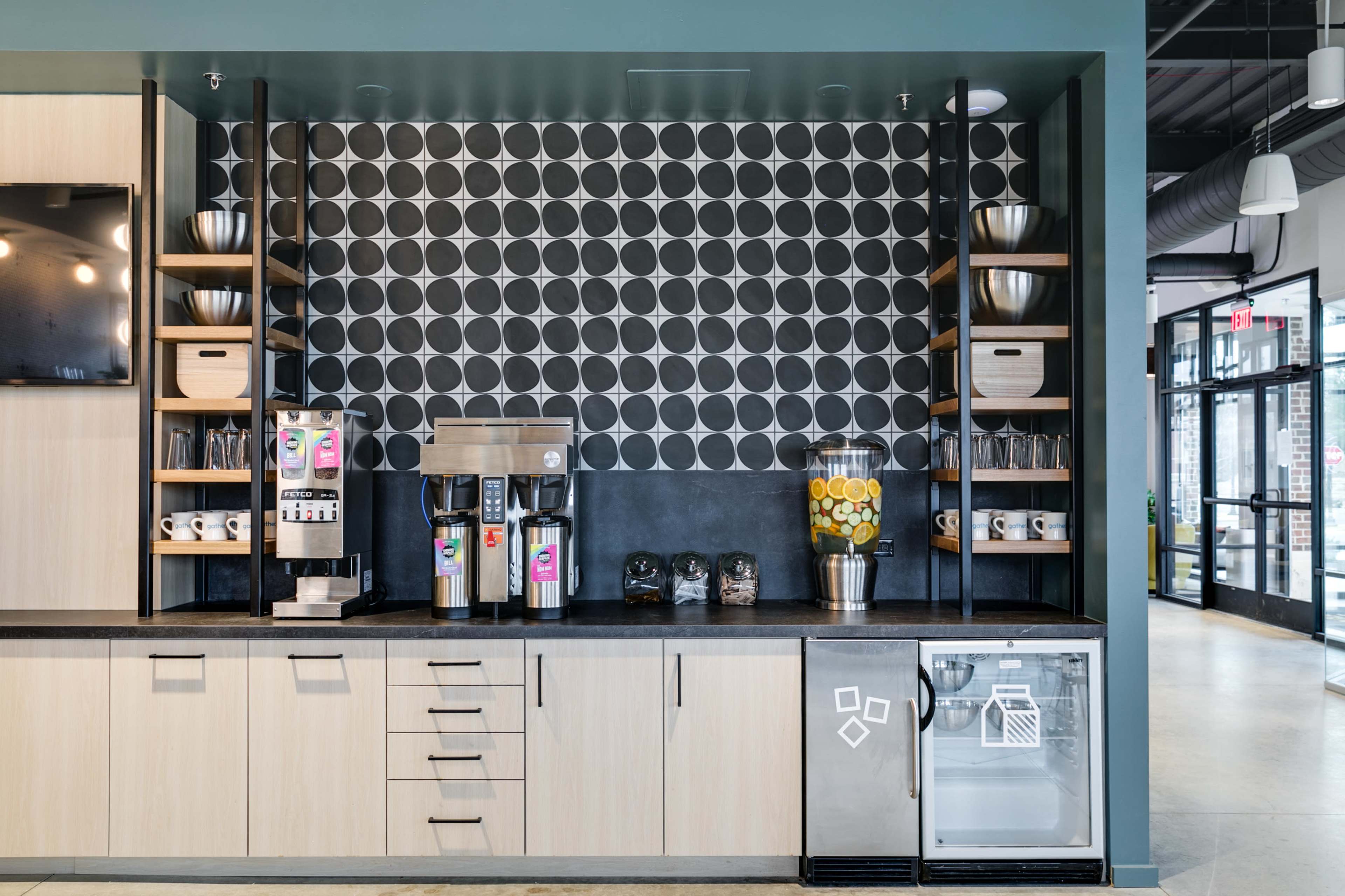 The image features a modern coffee station with a black and white circular patterned wall, a coffee machine, a display of citrus fruits, and a mini refrigerator.