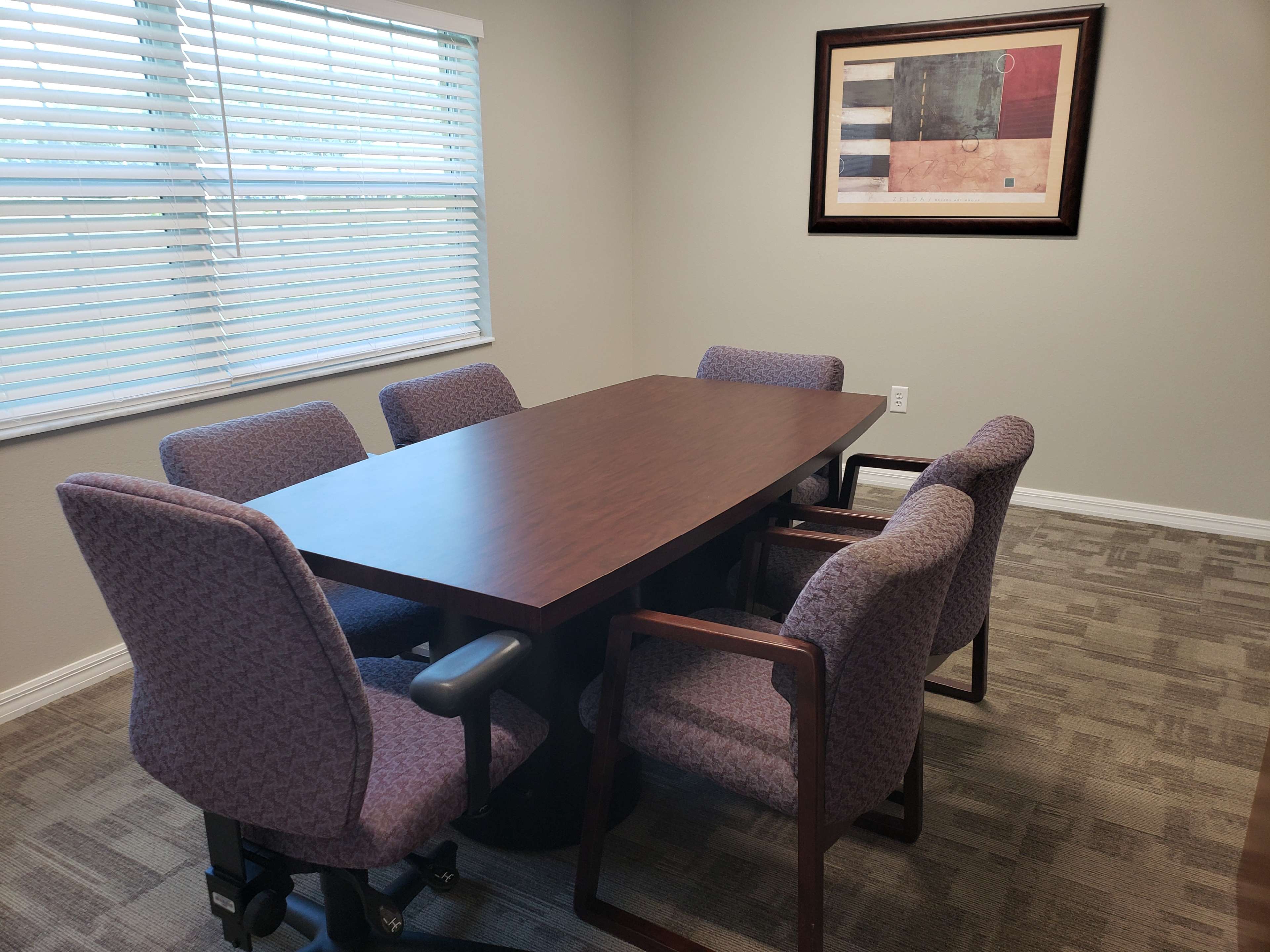 A conference room is set up with a large rectangular table surrounded by six upholstered chairs, and a framed piece of artwork hangs on the wall.