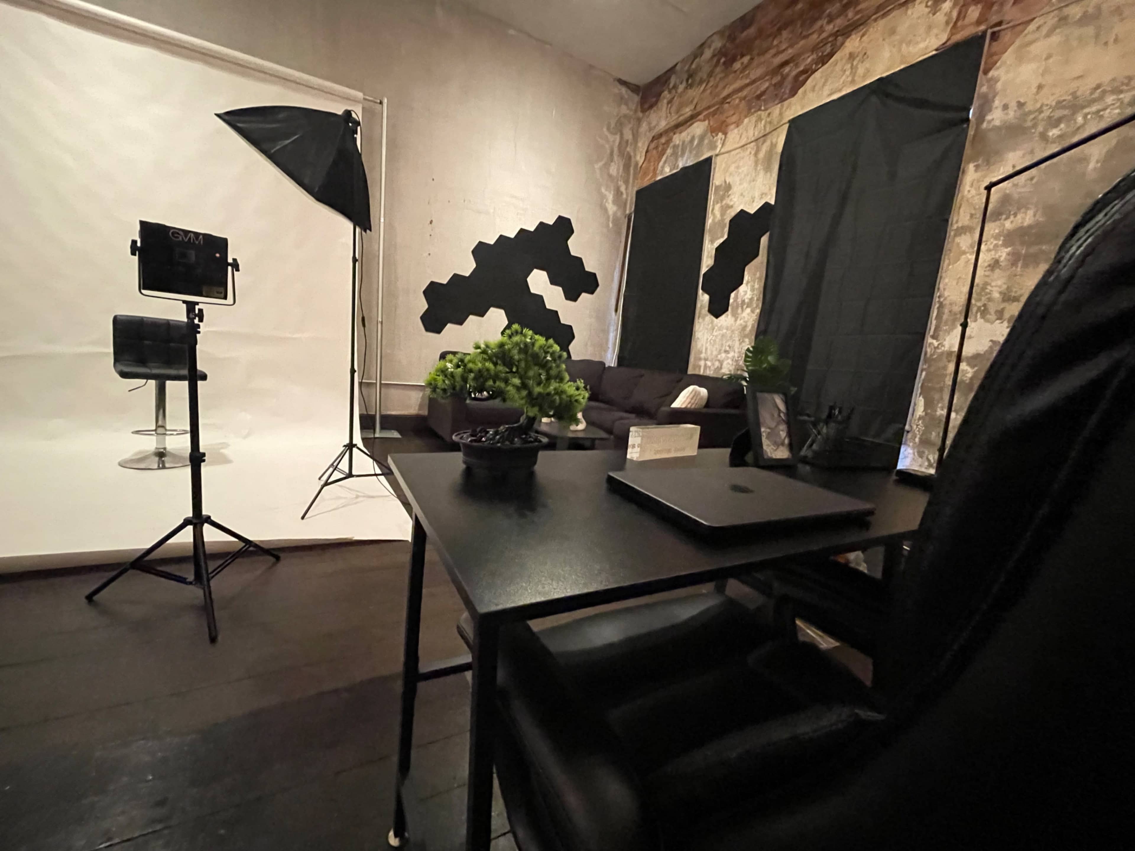 The image shows a photography studio with a black desk and chair, a light setup, a backdrop, and decorative plants in a room featuring textured walls and dark curtains.