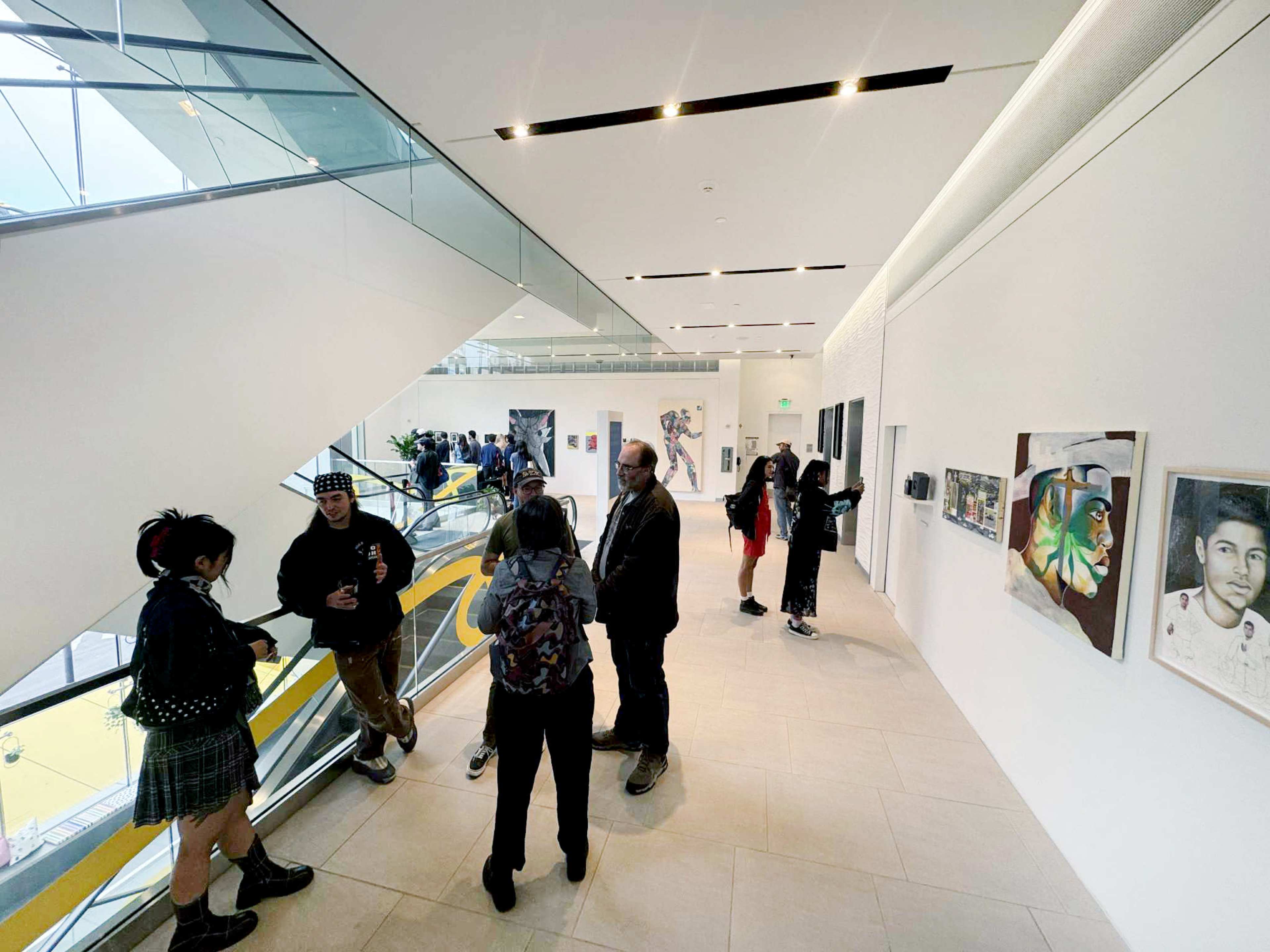 A group of people stands and chats in a bright gallery space featuring artwork on the walls and a staircase in the background.
