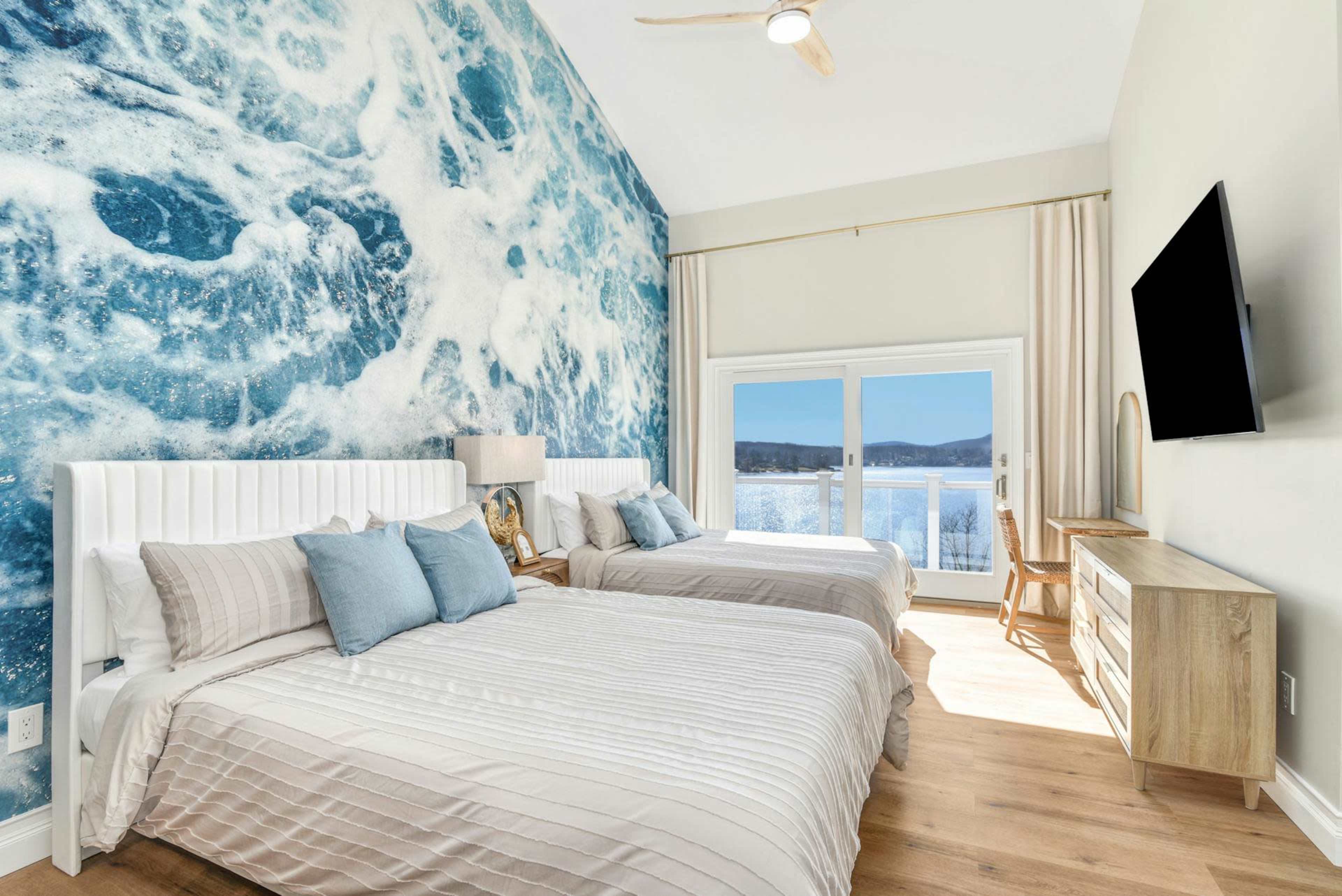 The image shows a bright bedroom with two beds, a large ocean wave mural on the wall, and a view of water through a sliding glass door.