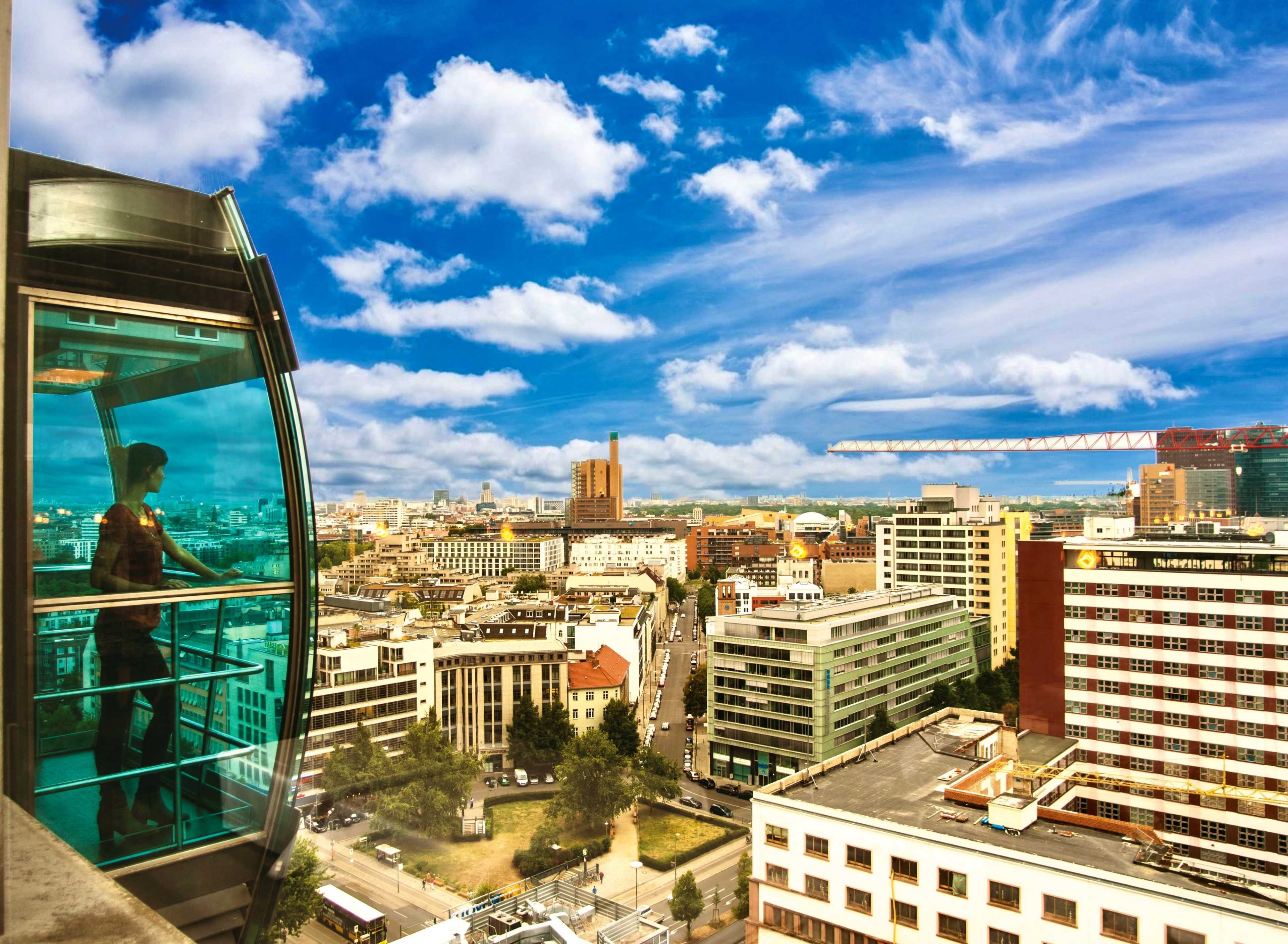 A person stands in a glass elevator overlooking a cityscape with a blue sky and scattered clouds.