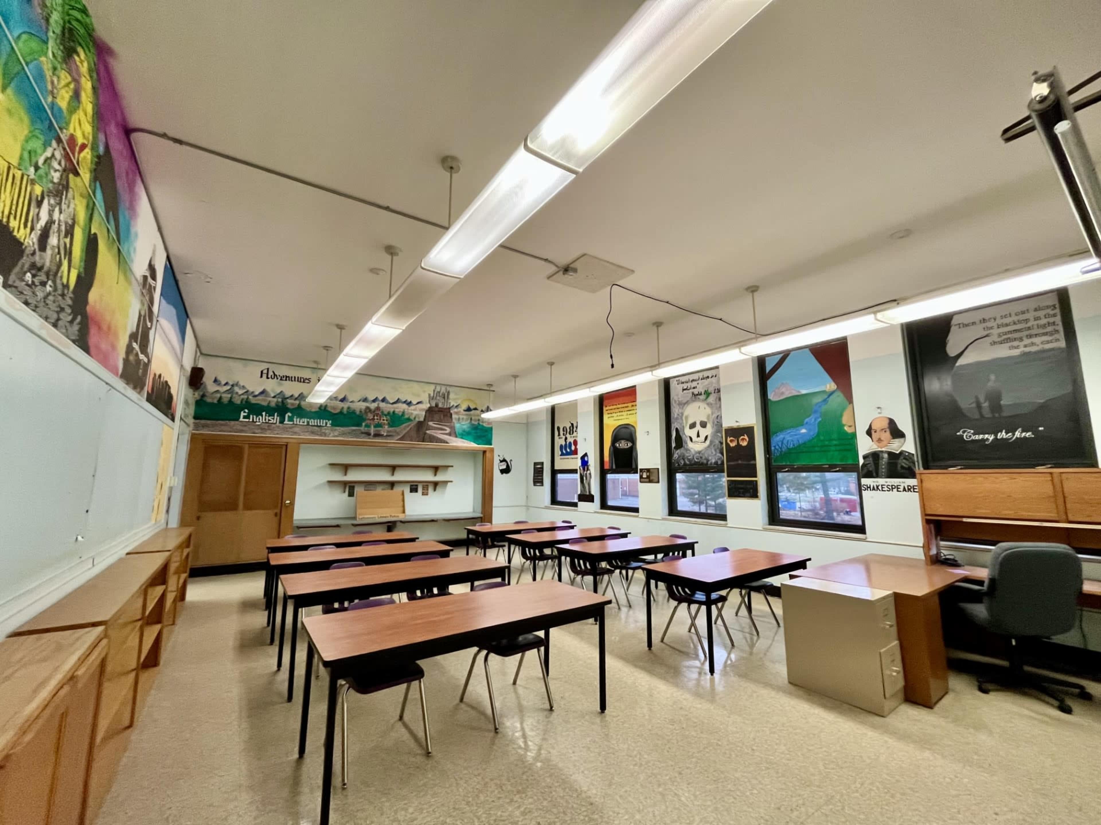 The classroom features several wooden tables arranged in rows, with colorful educational posters and murals decorating the walls.