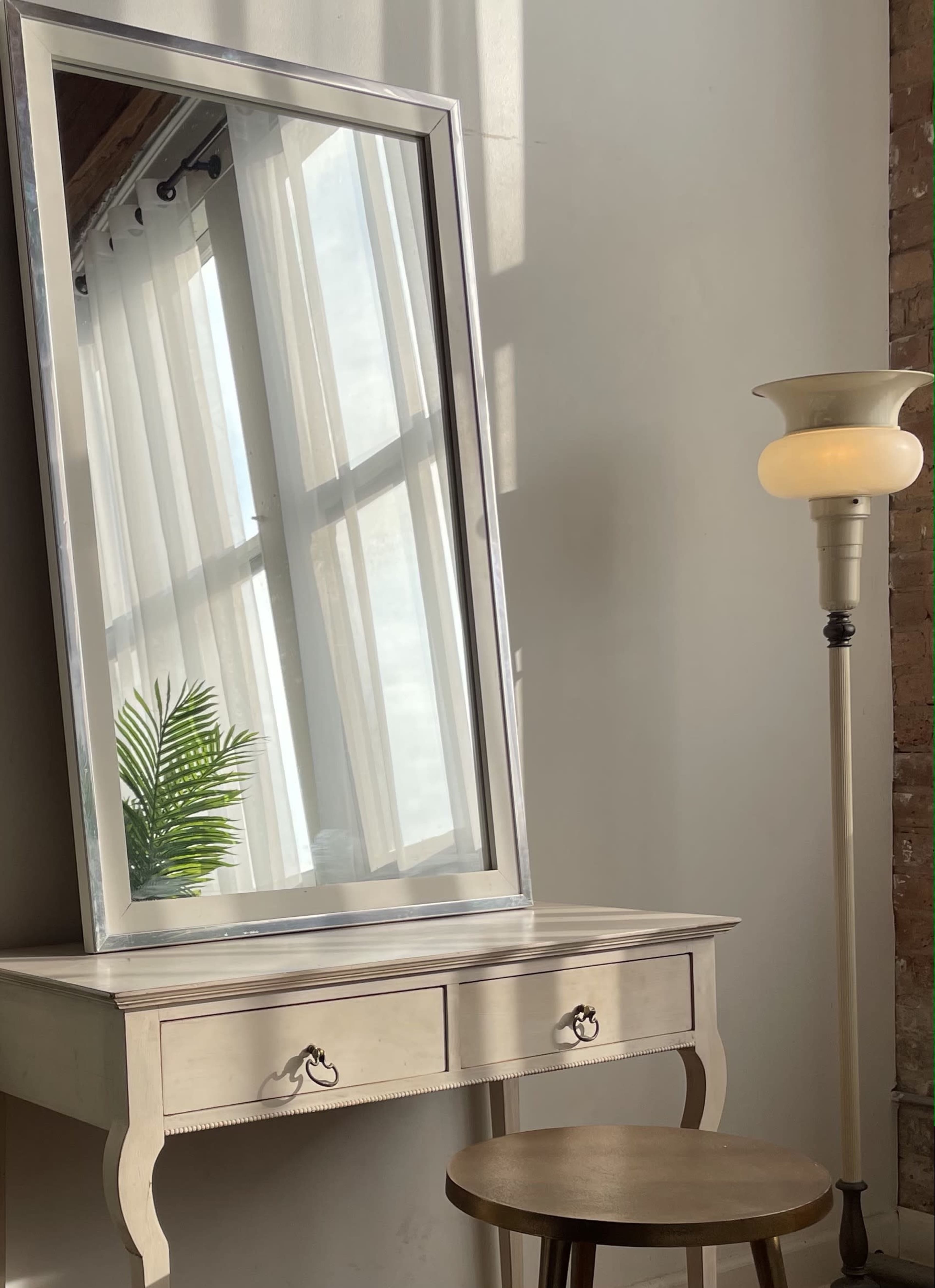 A large, framed mirror leans against a wall beside a small table with drawers and a lamp, while sunlight filters through sheer curtains.