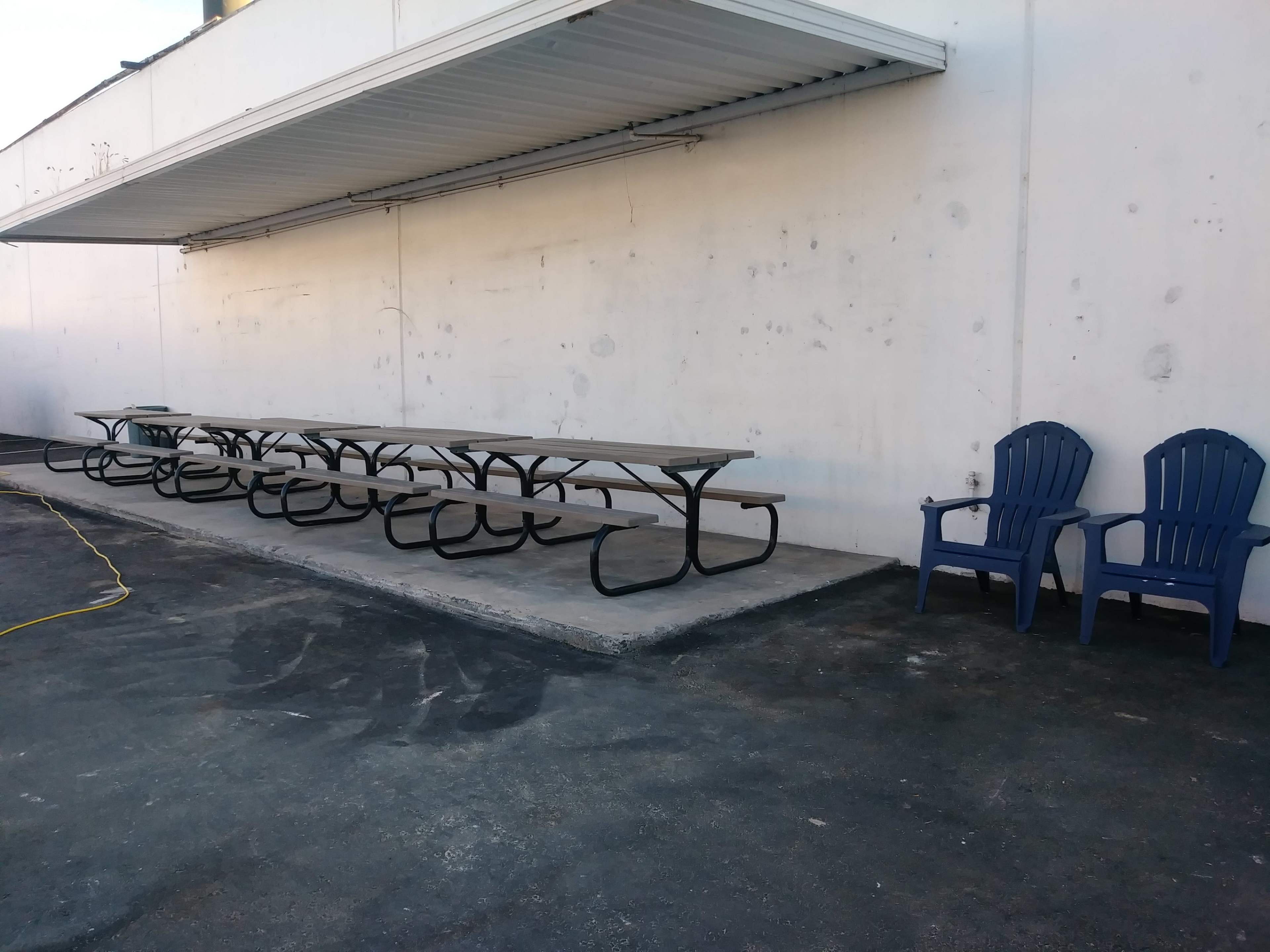 The image shows a concrete patio area with a long picnic table and a pair of blue lawn chairs against a white wall.