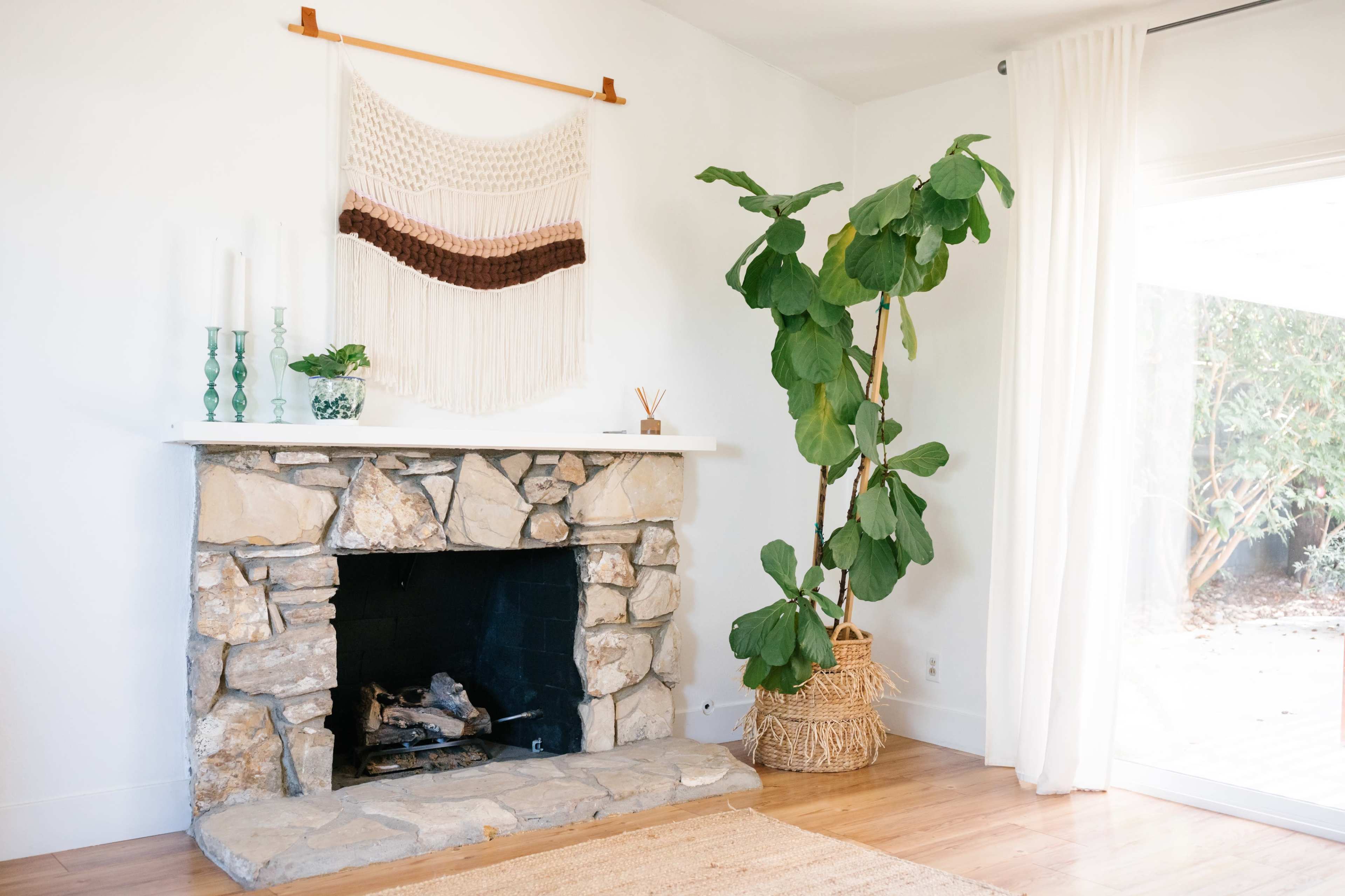 A stone fireplace is positioned against a white wall, decorated with a macramé wall hanging, and accompanied by a tall leafy plant in a woven basket.