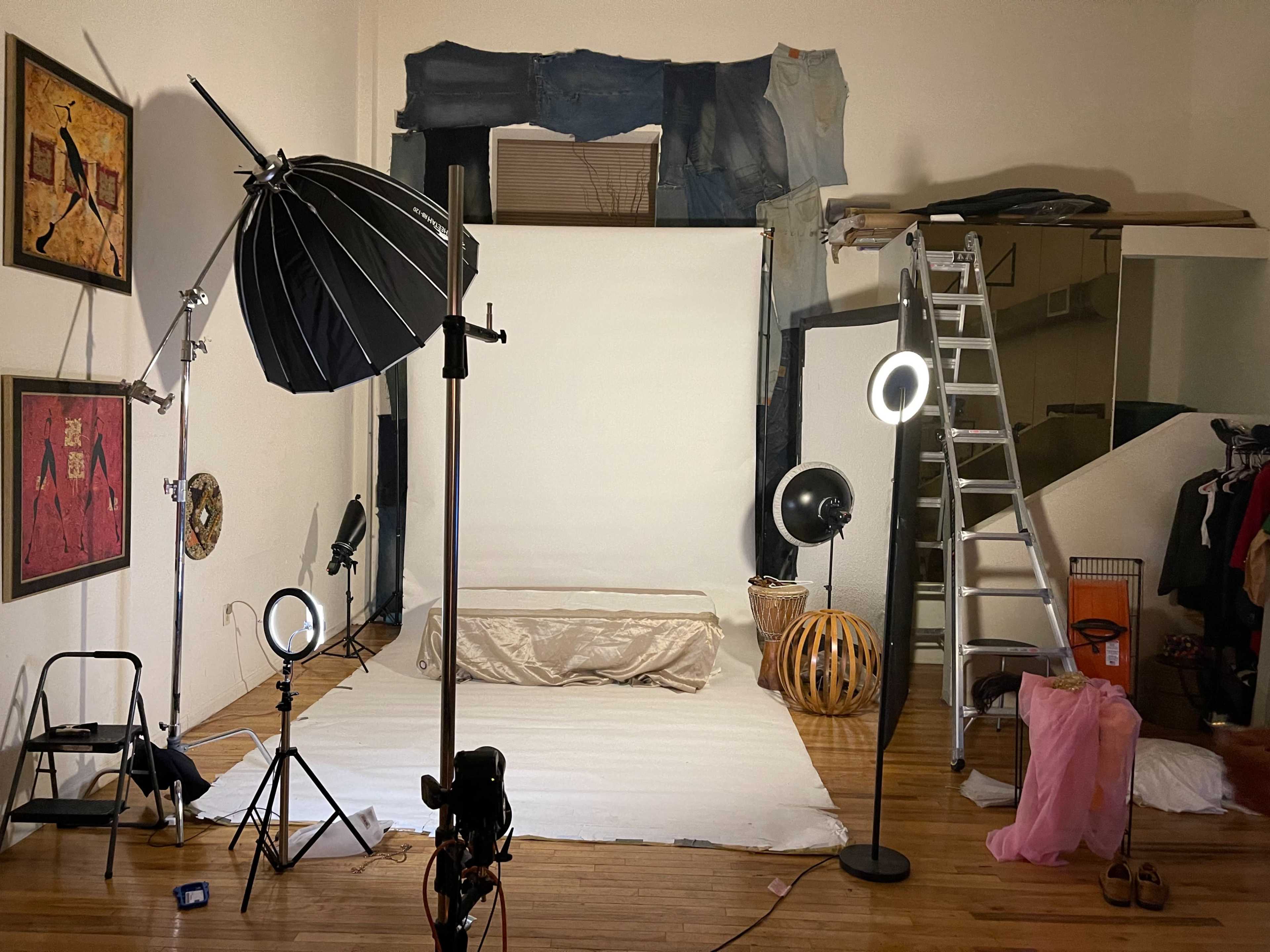 The image shows a photo studio setup with a white backdrop, lighting equipment, a ladder, and various props scattered around the floor.