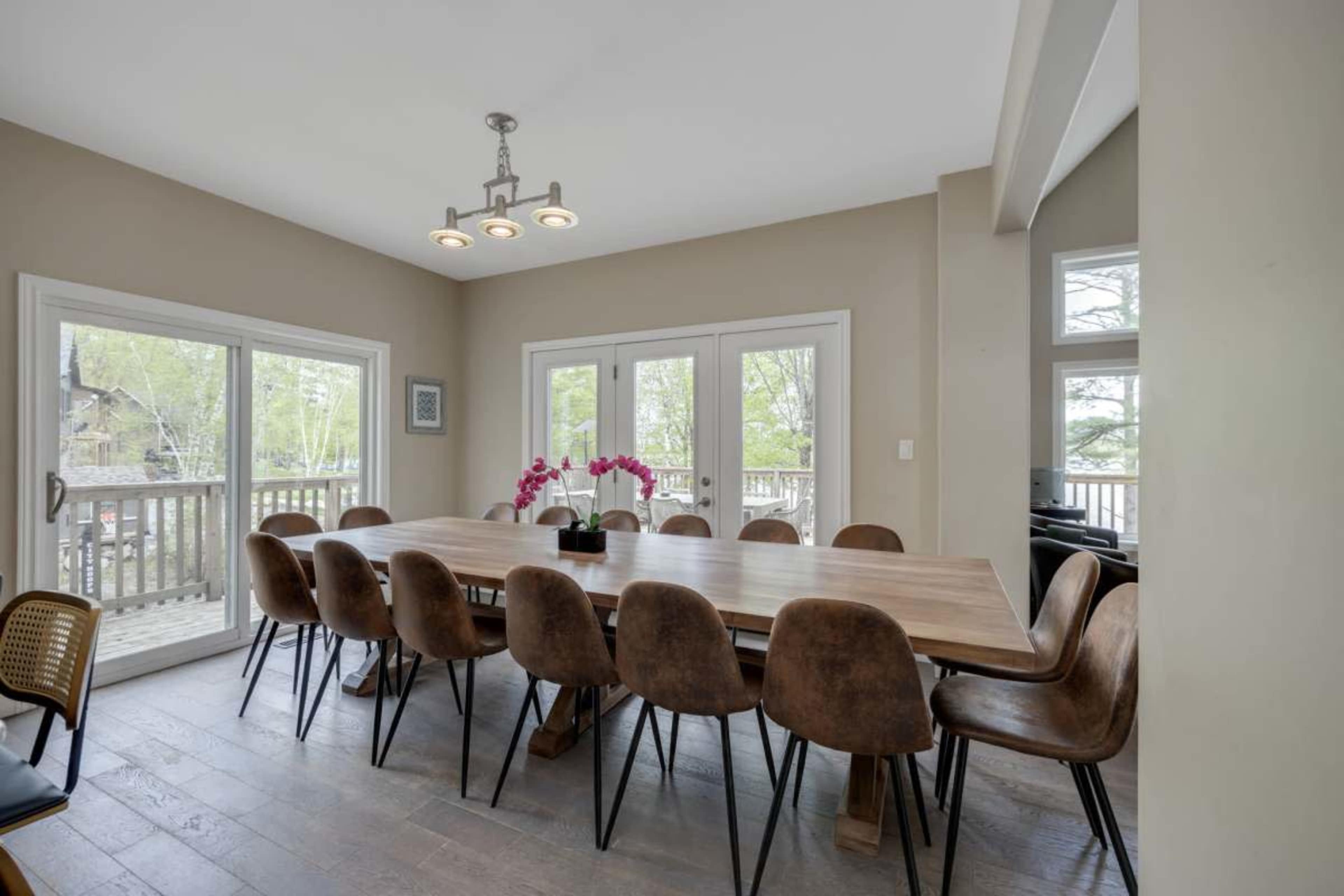 A spacious dining area features a long wooden table surrounded by brown chairs, with large windows overlooking a deck and greenery outside.