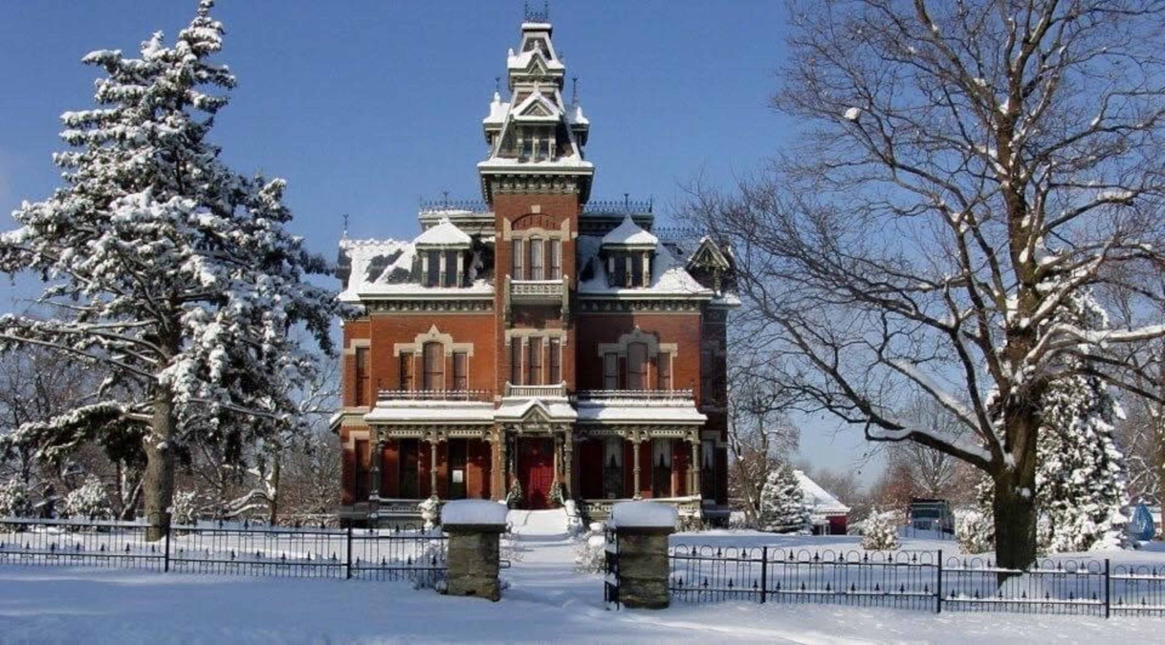 A large Victorian mansion with ornate architecture is surrounded by snow-covered trees and a white landscape in winter.