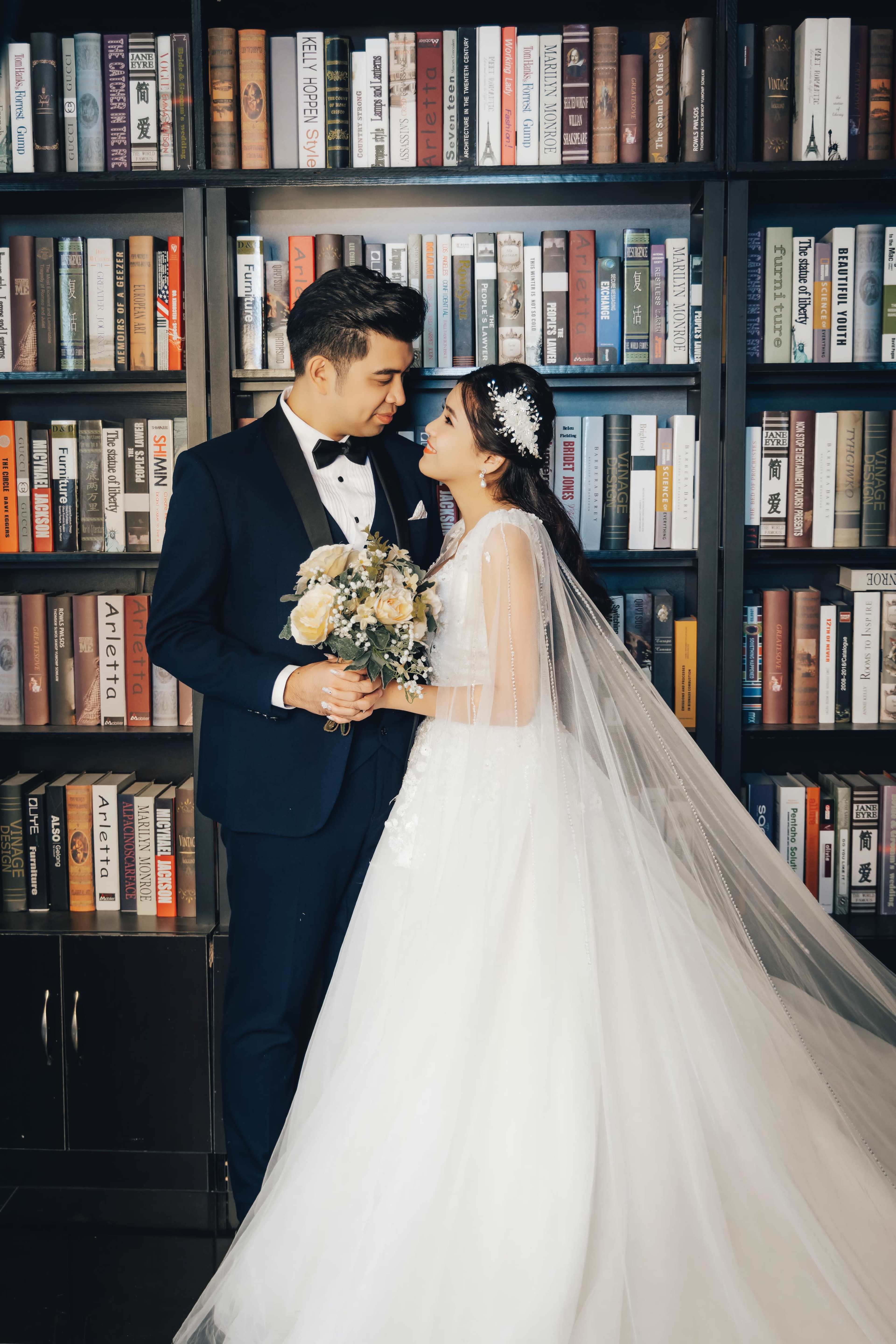 A bride and groom stand together in a library, smiling at each other while holding a bouquet of flowers.