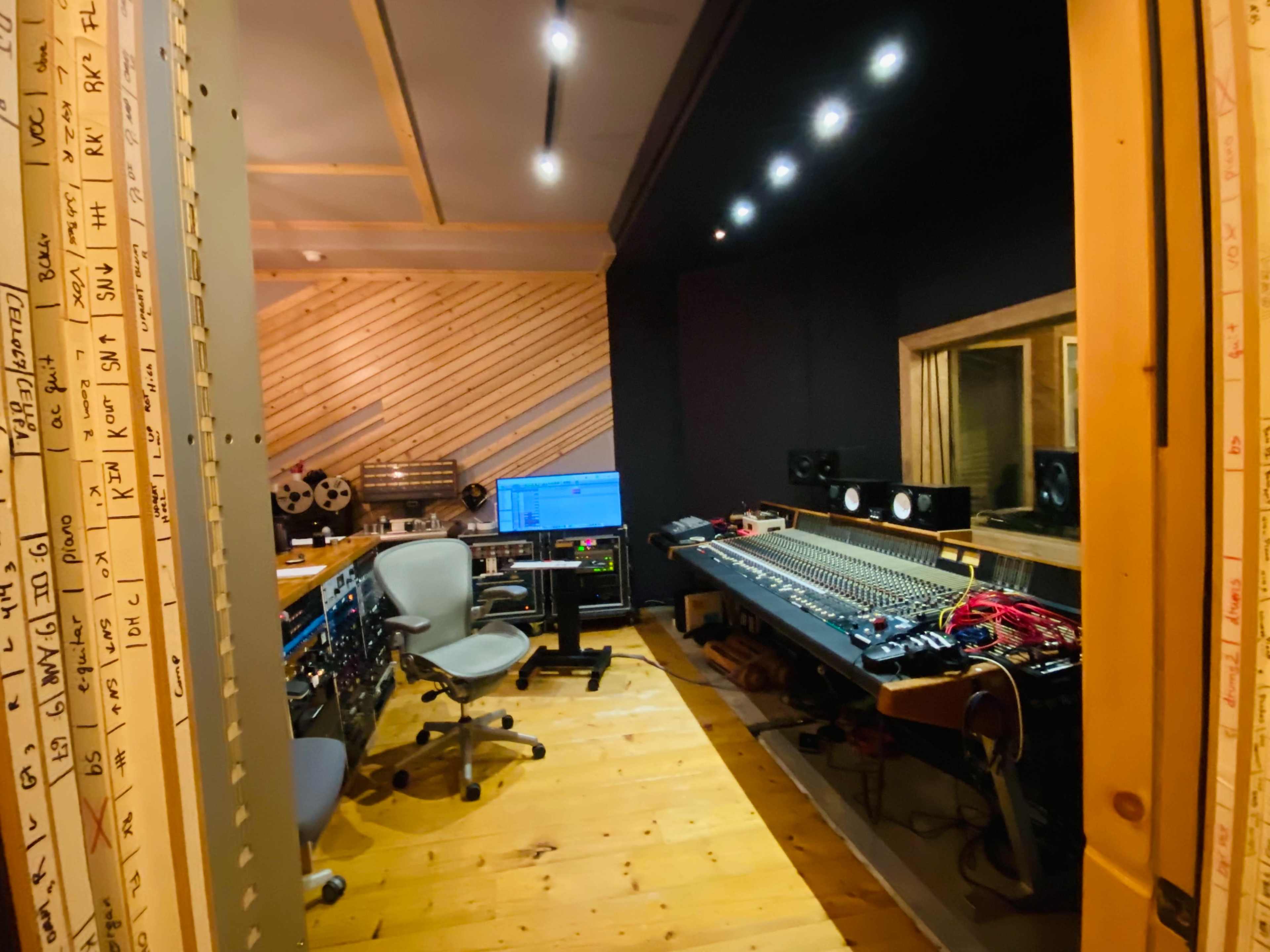 A recording studio with a mixing console, computer setup, and various audio equipment along wooden walls and a wooden floor.