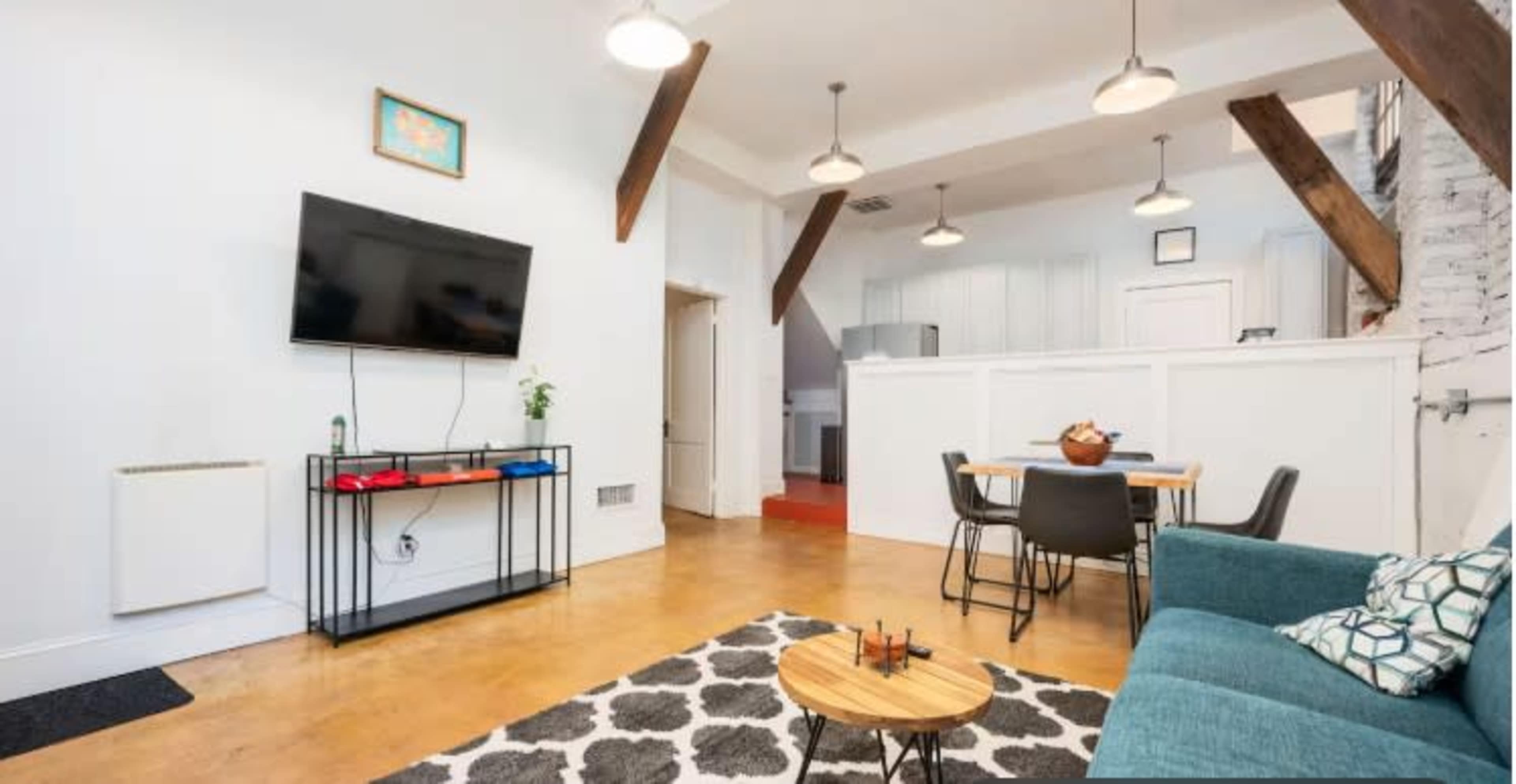 The image shows a modern living space featuring a television mounted on the wall, a blue sofa, a dining area with a wooden table and chairs, and exposed wooden beams on the ceiling.