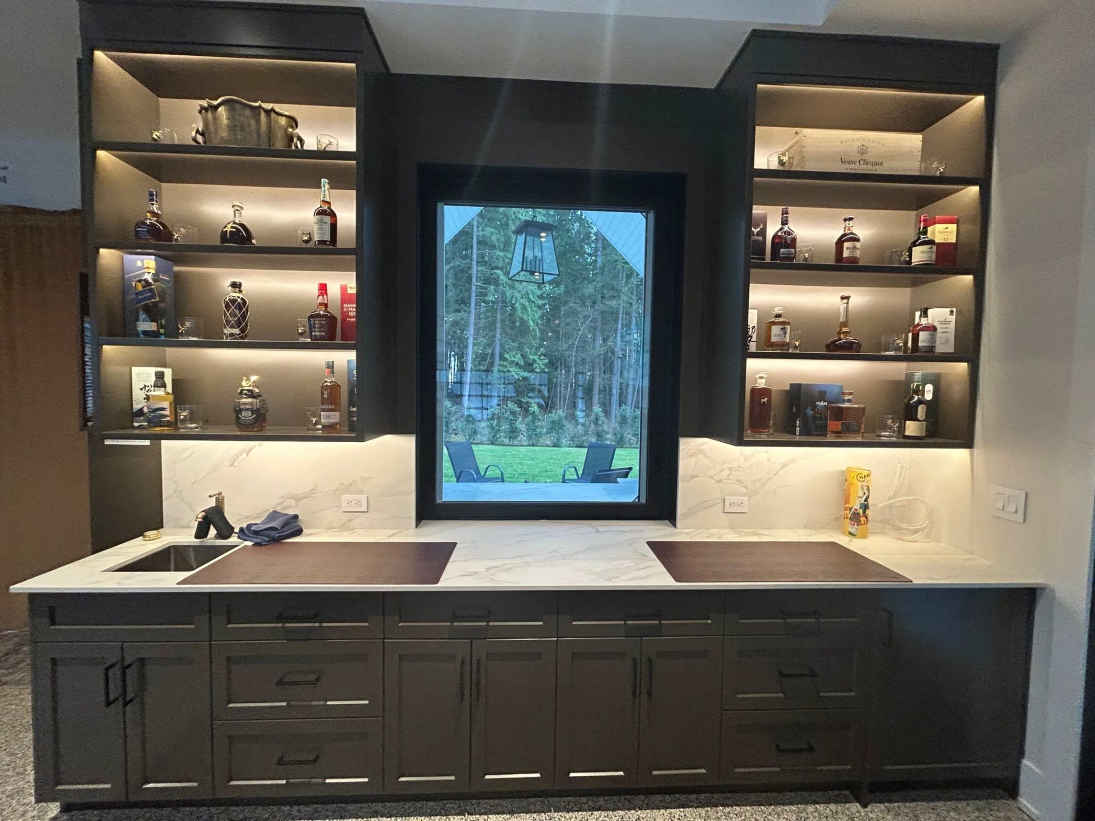 The image shows a neatly designed bar area with a marble countertop, dark cabinetry, and backlit shelves displaying various bottles of liquor, framed by a large window overlooking greenery.