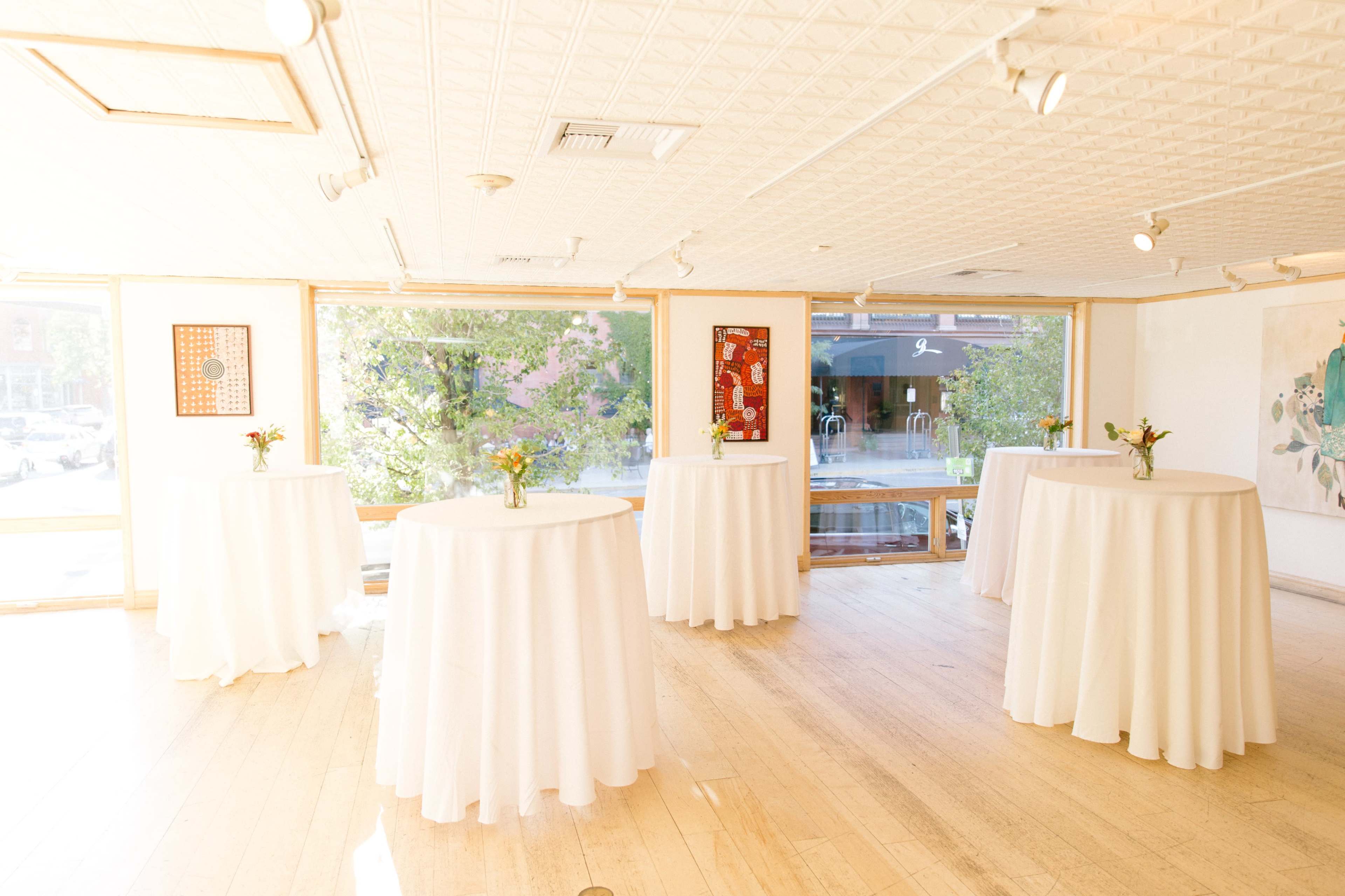 The image shows a bright gallery space featuring several round tables dressed in white tablecloths, with windows overlooking a tree-lined street.