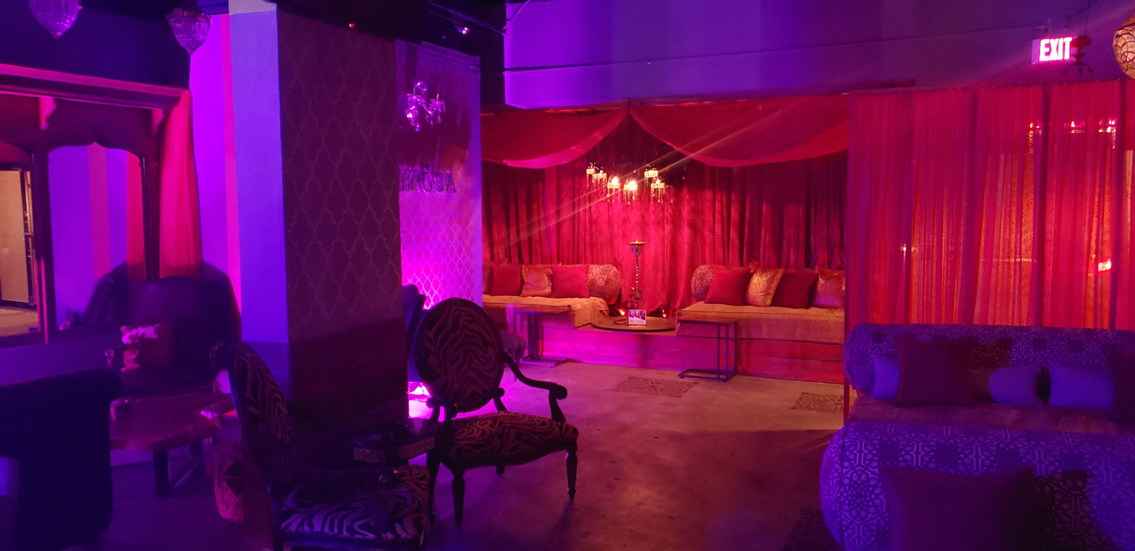 An interior space with plush couches, patterned chairs, and purple lighting, separated by sheer curtains.