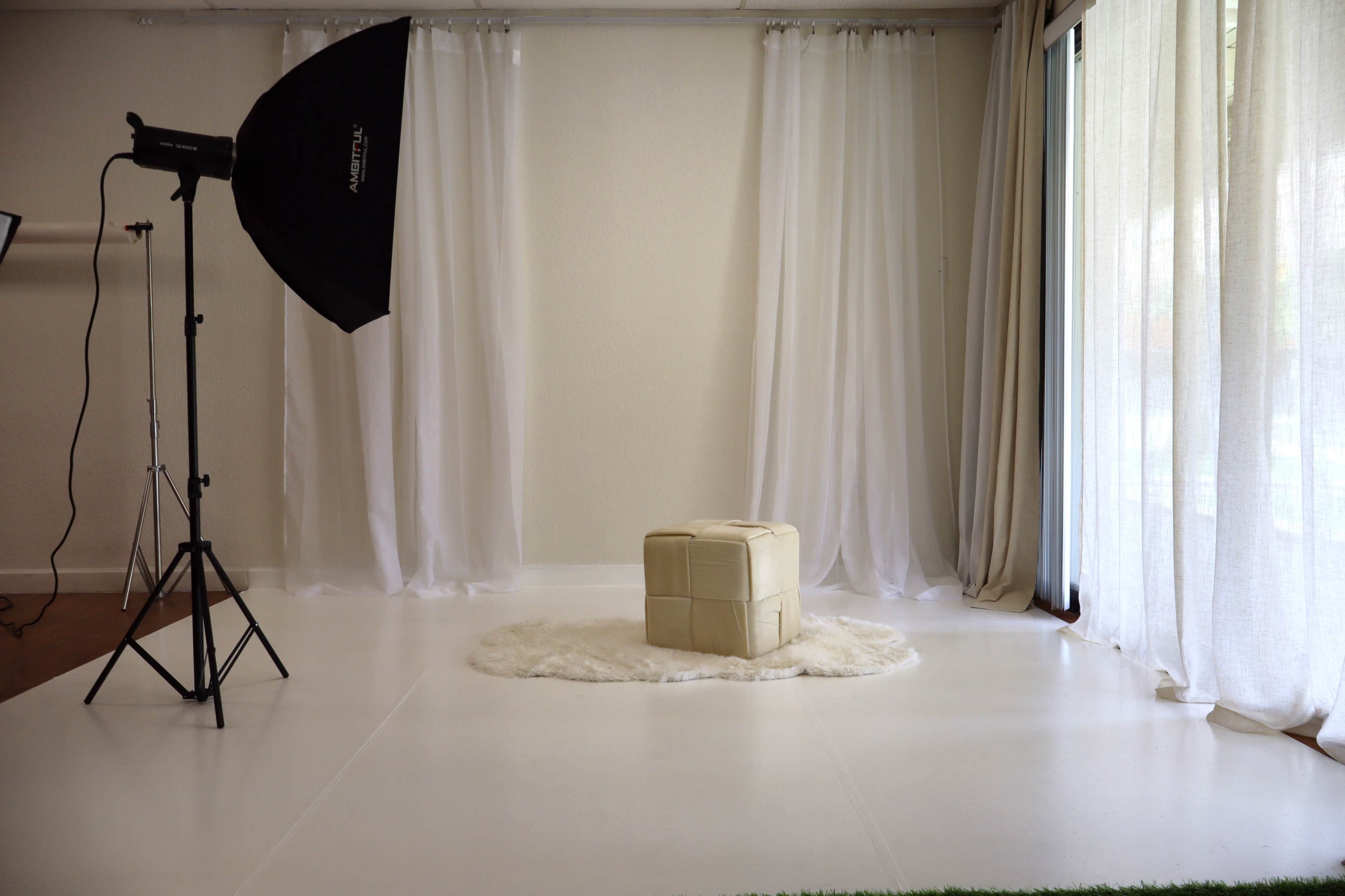 A photography studio features a soft-lit background with sheer curtains, a light stand, and a small, upholstered cube on a circular rug.