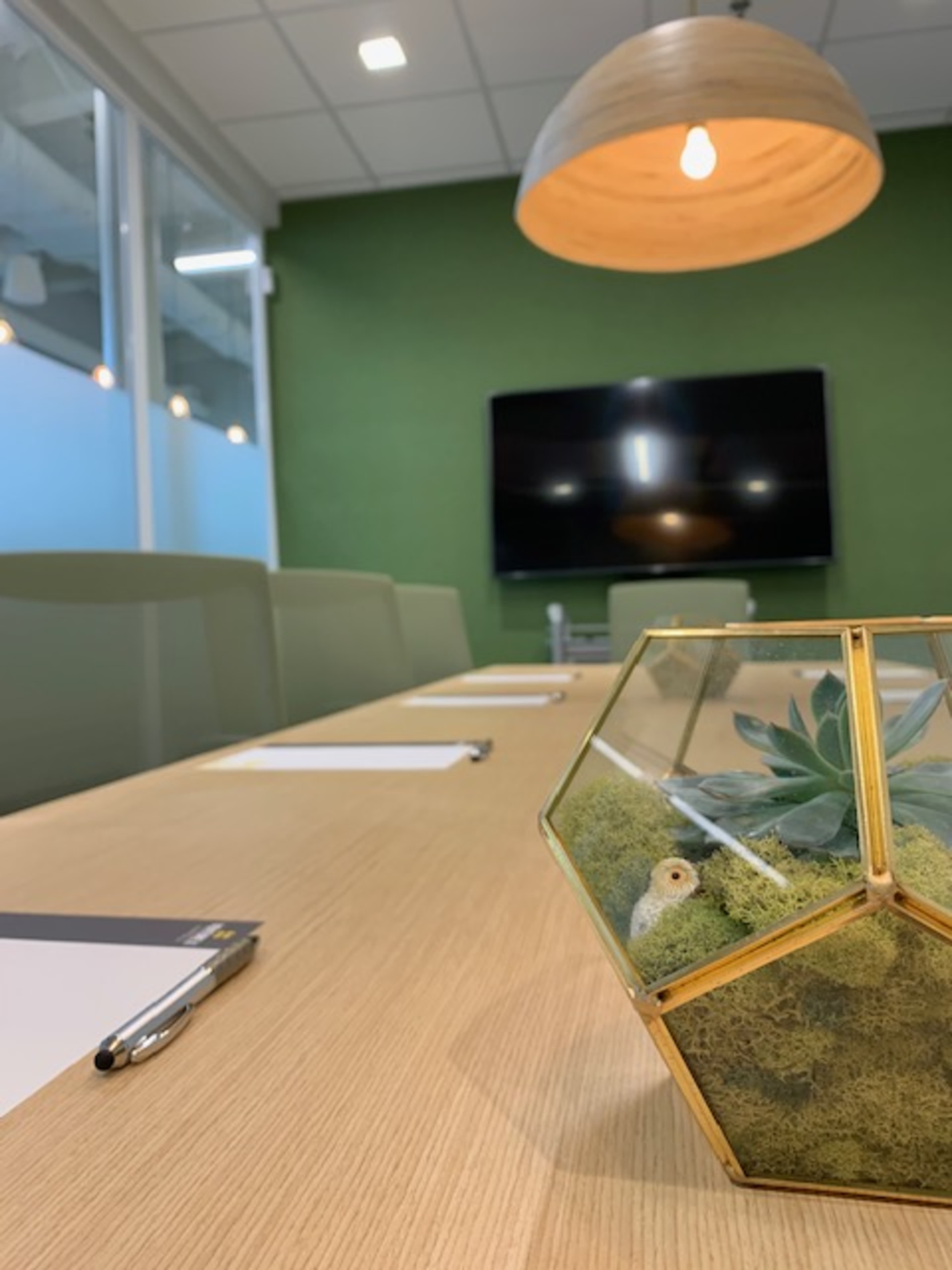 A conference room table is set with notepads and pens, featuring a geometric glass planter with succulents and a hanging light fixture above.