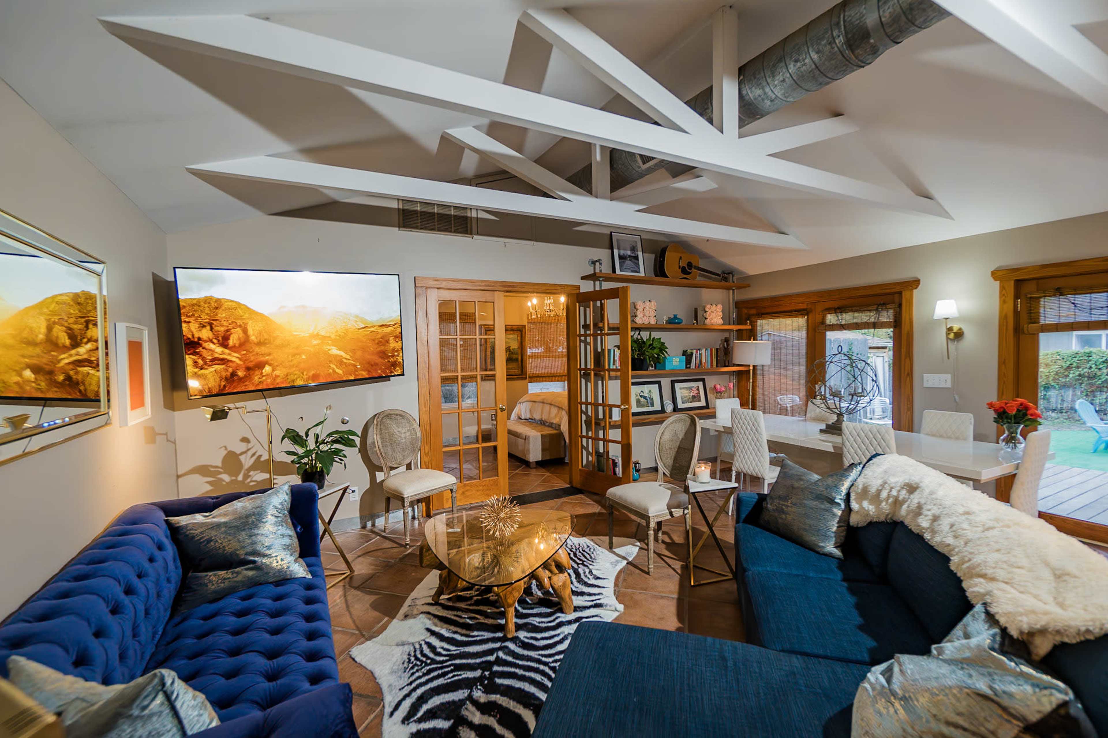 The image shows a modern living space with a blue sectional sofa, a glass coffee table, and a large television mounted on the wall, featuring a wooden bookshelf and dining area in the background.